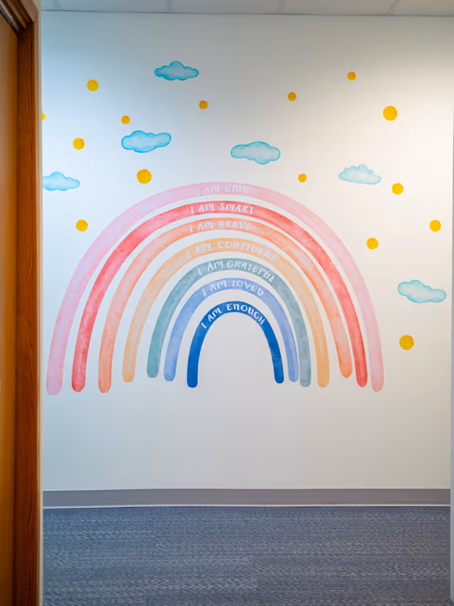 Colorful rainbow mural with clouds on a white interior wall in a hallway.