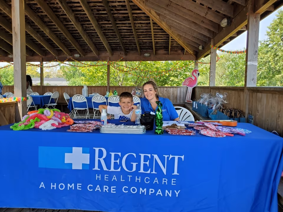 Two smiling people sit at a blue Regent Healthcare table at an outdoor pavilion.