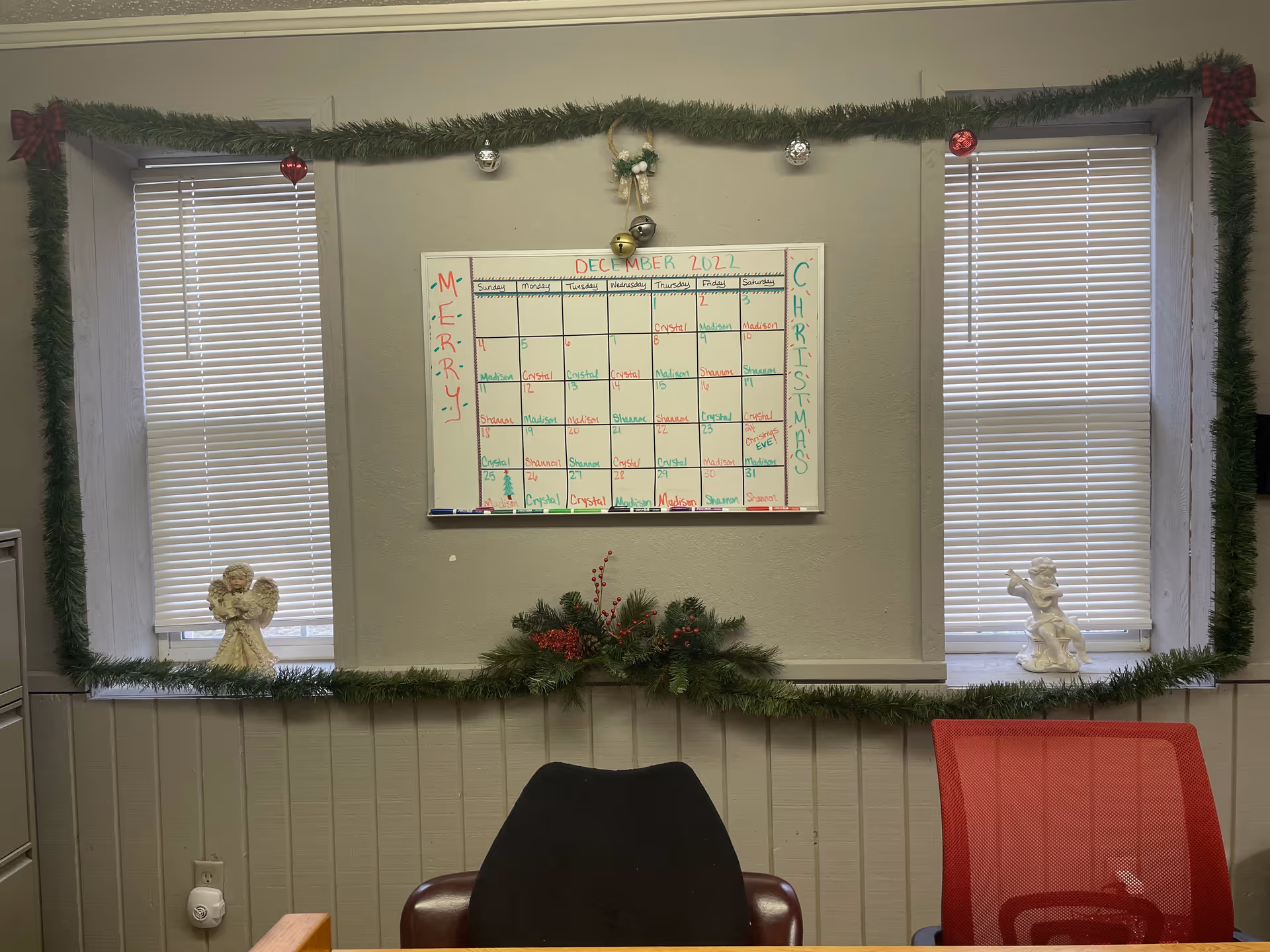 Office area with calendar on wall, festive garlands, two windows, and chairs.