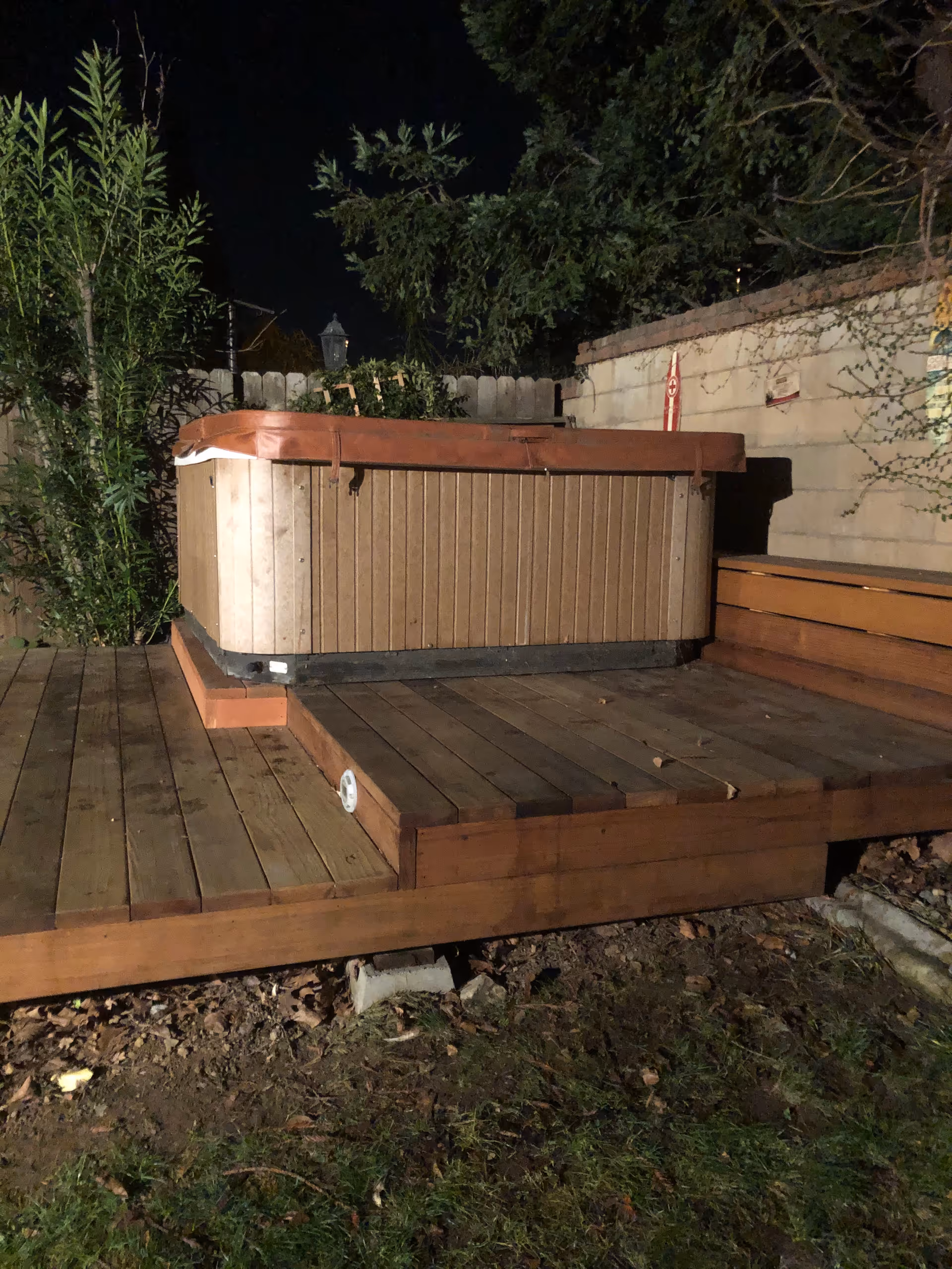 Nighttime view of a wooden outdoor deck with a hot tub cover and surrounding greenery.