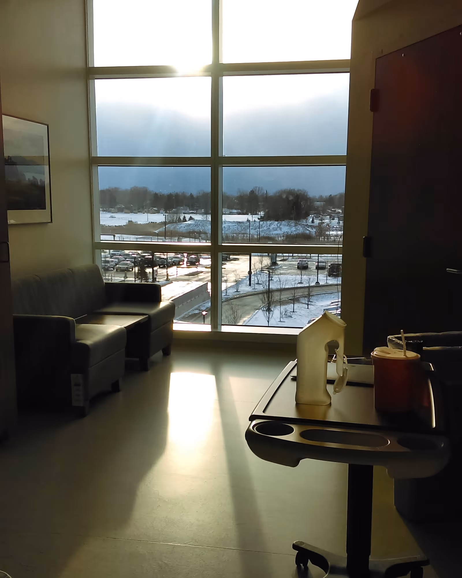 Sunlit hospital lobby with a large window view, seating area, and a serving cart.