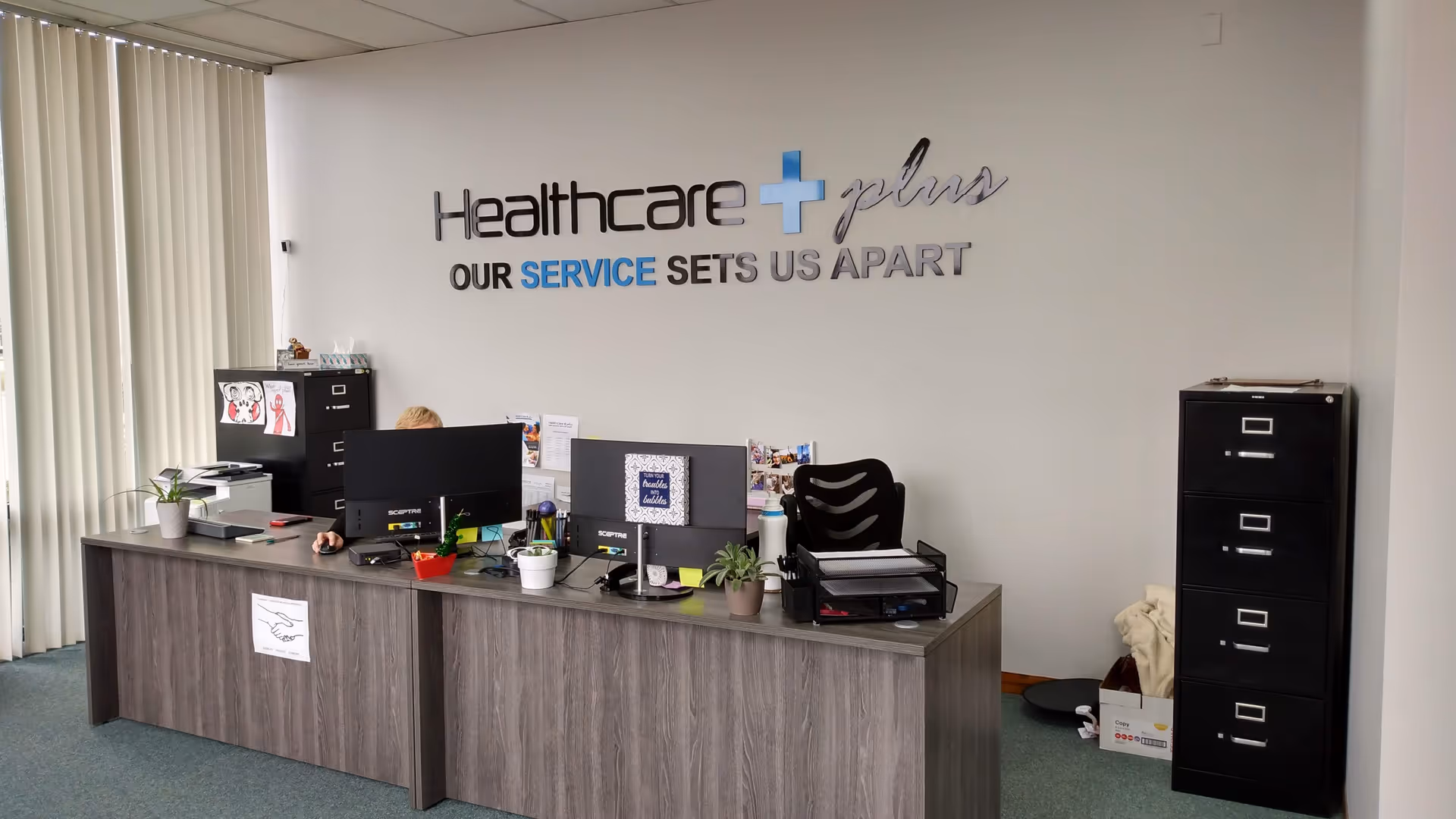 Reception desk in a healthcare office with computers, plants, and a Healthcare Plus sign
