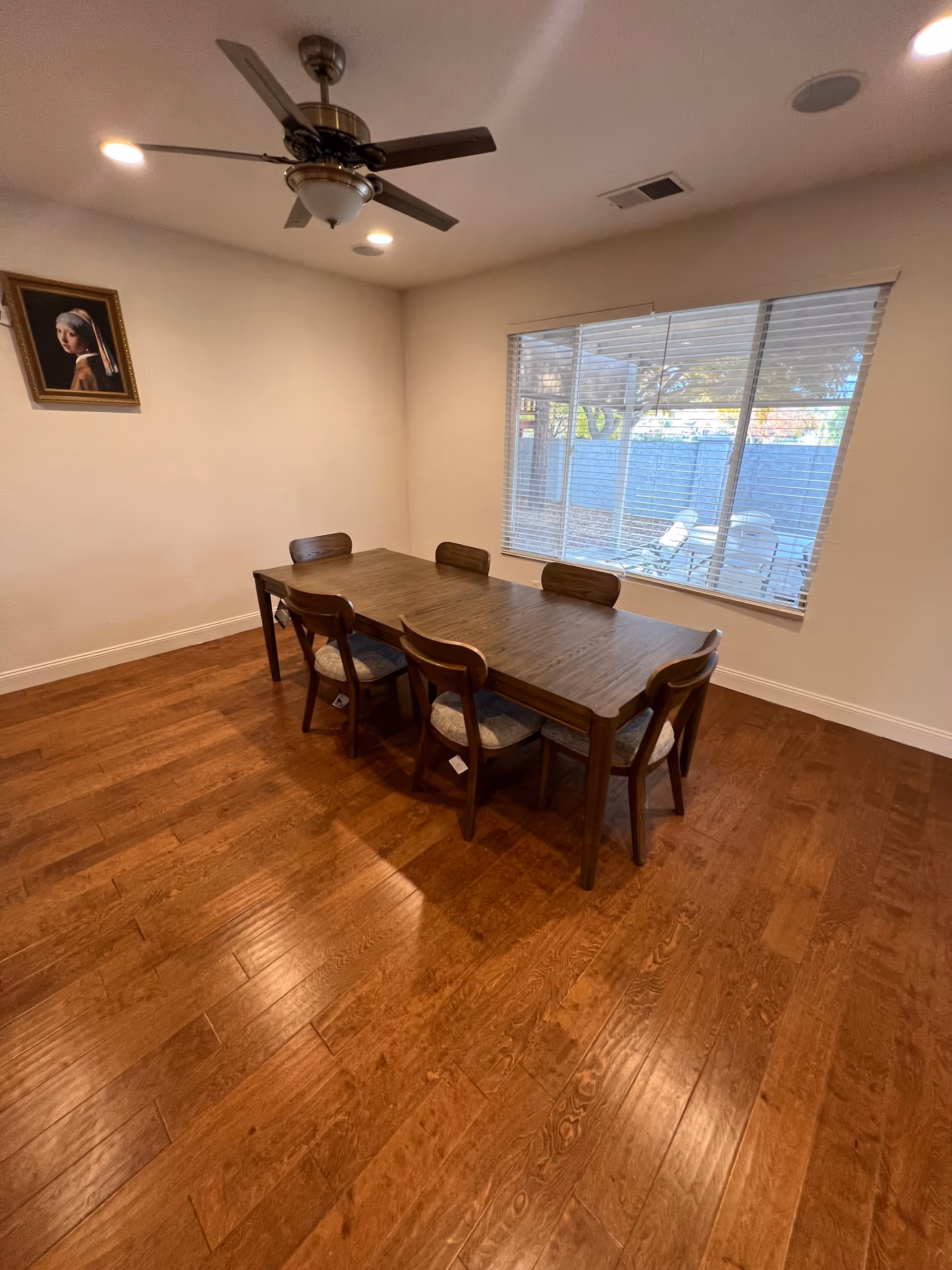 A dining room with a wooden table and several chairs by a window and ceiling fan.