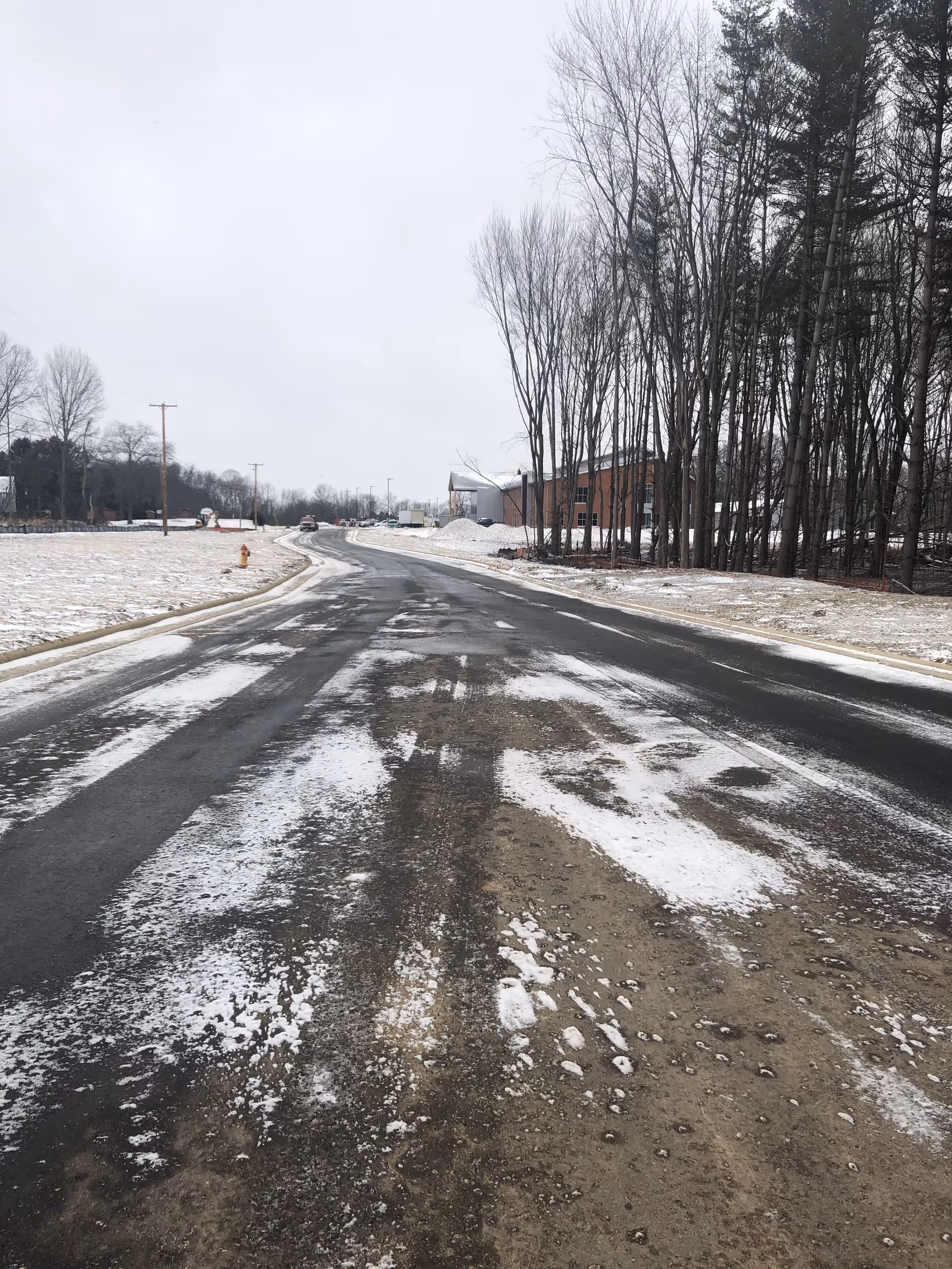 Snowy road beside leafless trees and a brick building under an overcast sky