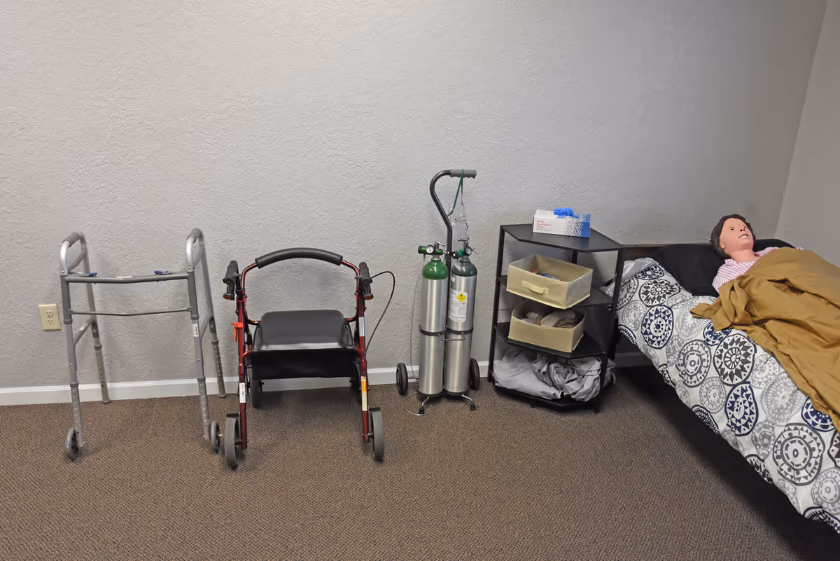 A small bedroom with bed, medical cart, oxygen tanks, walker, and a mannequin in bed.