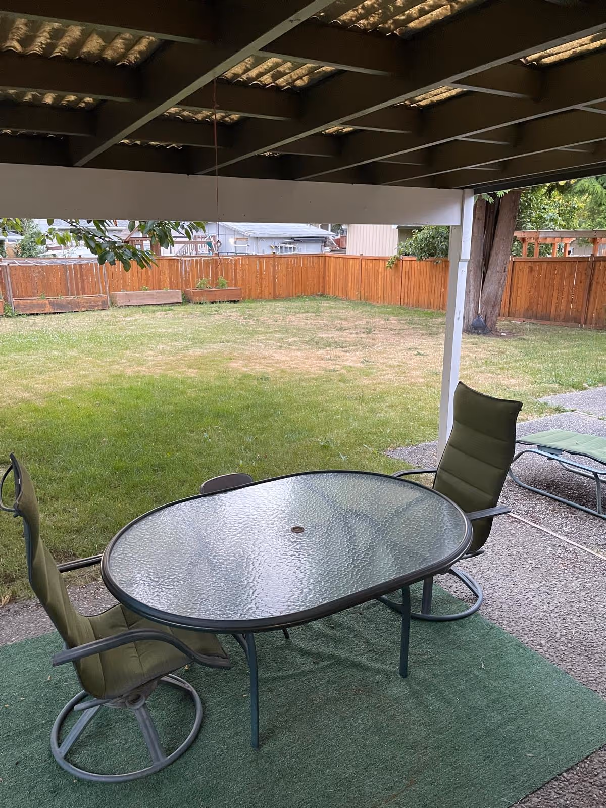 Under a covered patio, glass table with chairs overlooking a fenced grassy backyard.
