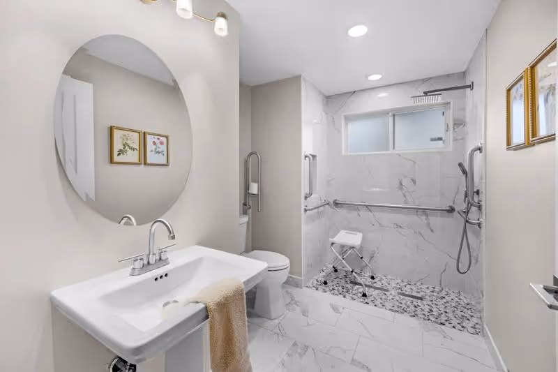 Accessible bathroom with sink, round mirror, toilet, and walk-in marble shower.