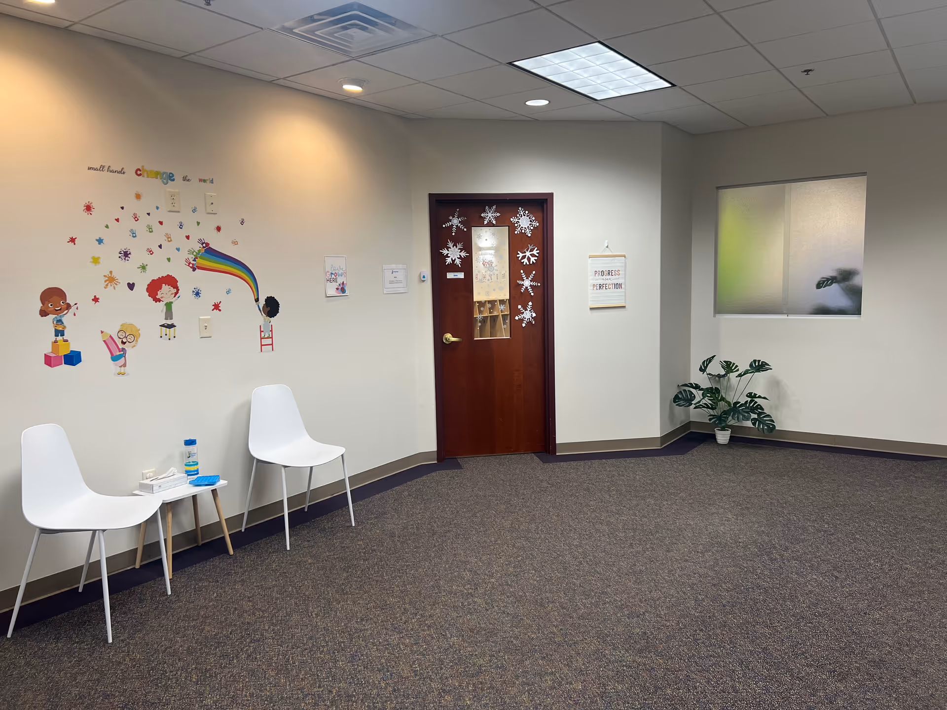 Interior waiting area with white chairs, wall decals, a wooden door, and a potted plant.