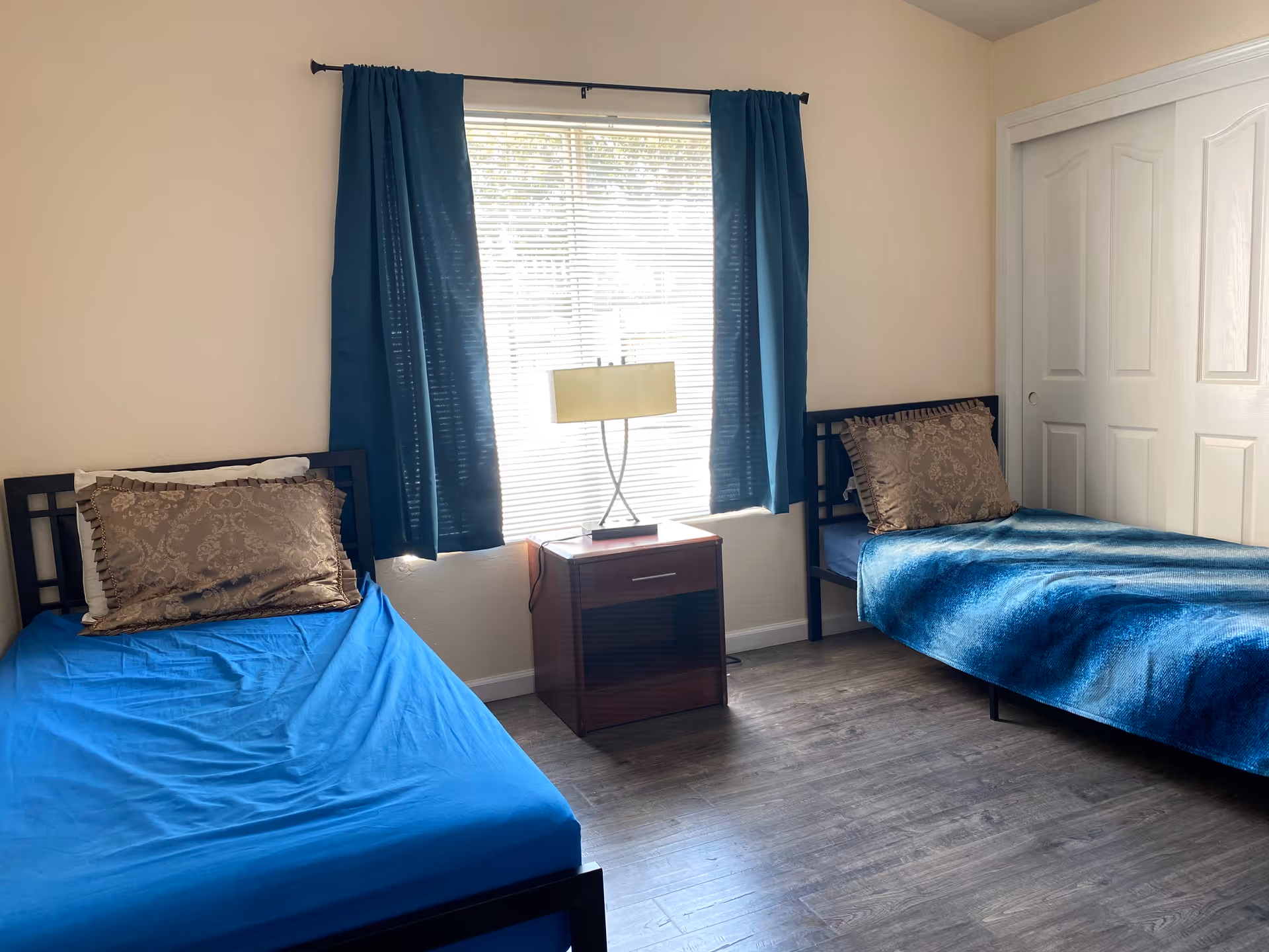 Two twin beds with blue bedding and a nightstand between a window with blue curtains.