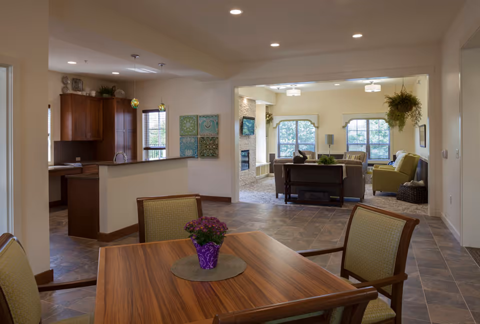 Open, well-lit senior living common area with dining table, kitchen counter, and seating.