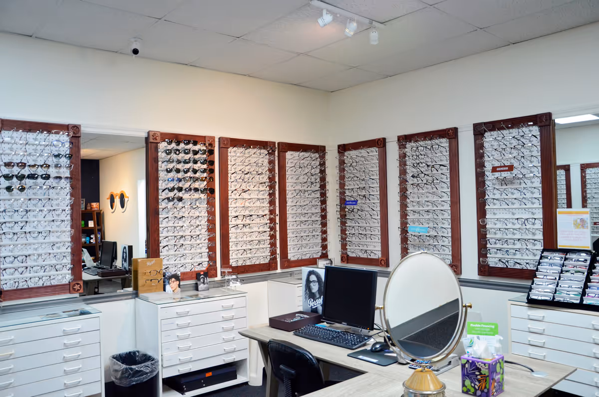 Eyewear shop interior with wall-mounted frame displays, a desk, computer, and round mirror.