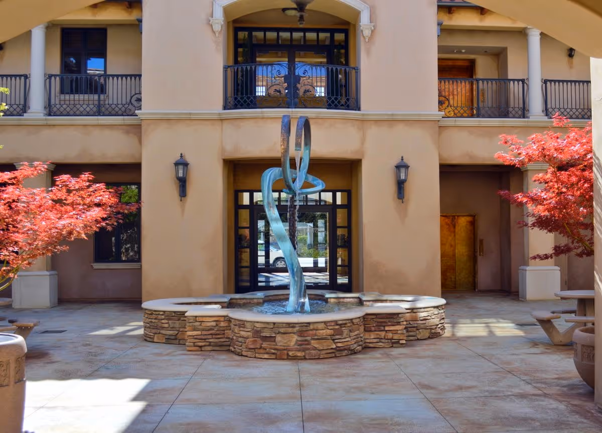 Exterior courtyard of a senior living facility featuring a sculptural fountain and red trees.