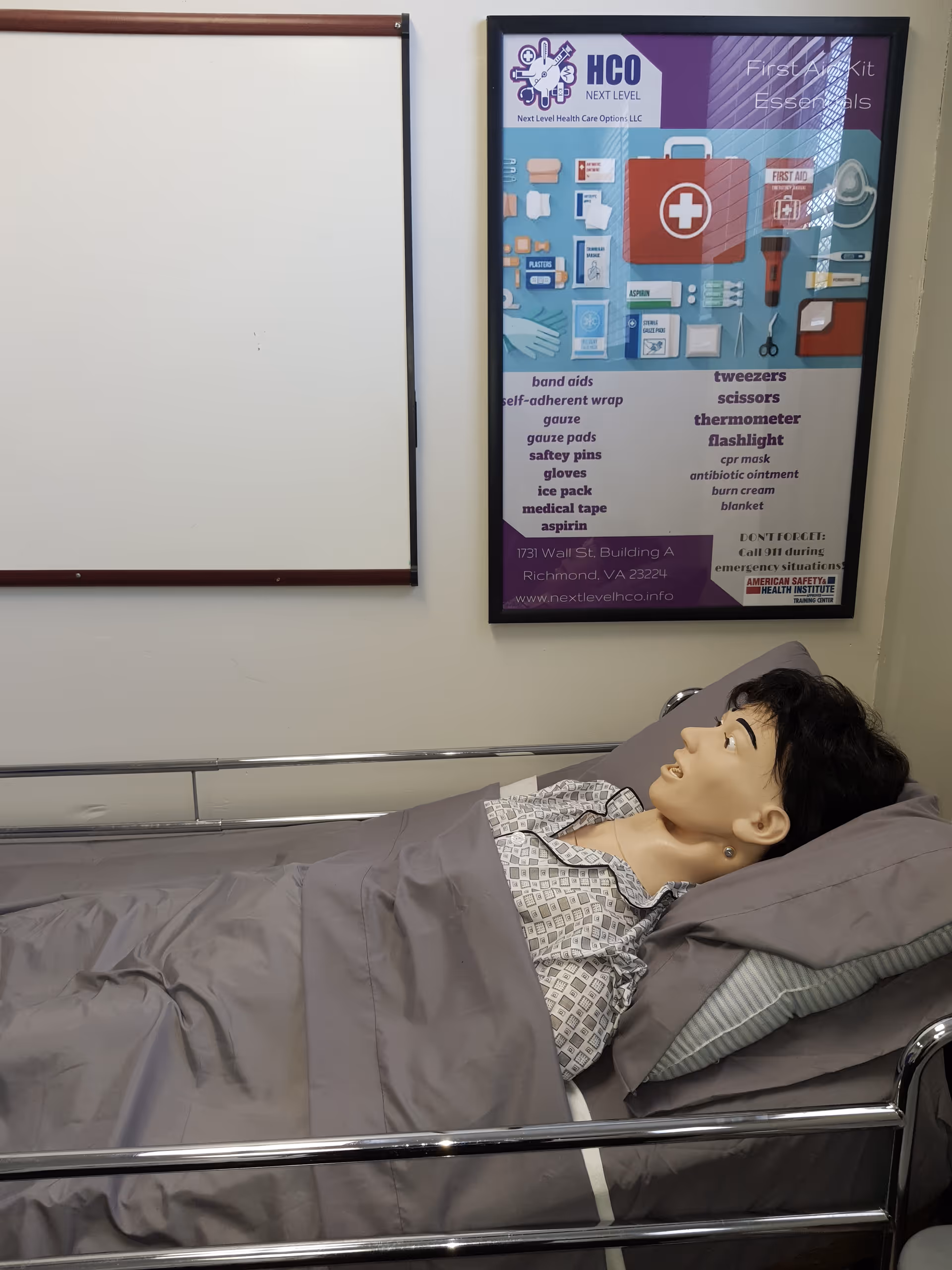 A medical training mannequin lies in a hospital-style bed in a patient room.
