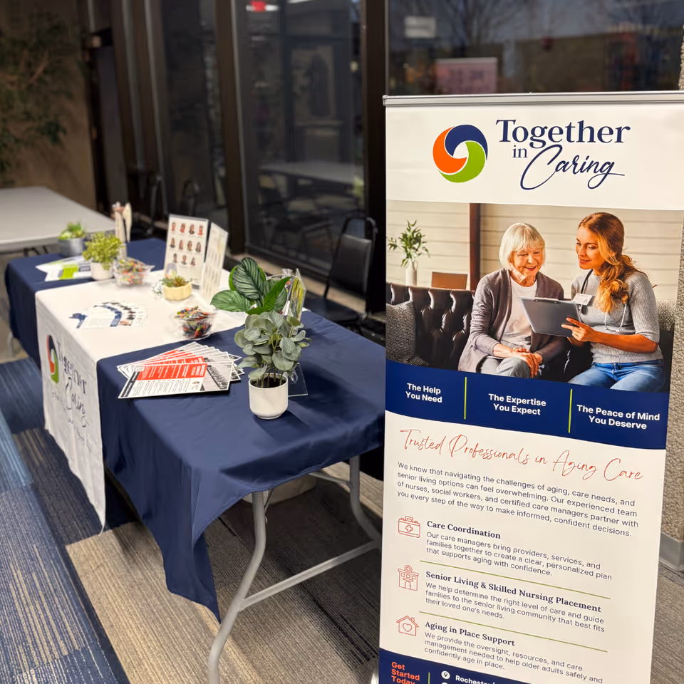 Indoor display table with navy cloth, brochures, plants, and a Together in Caring banner.