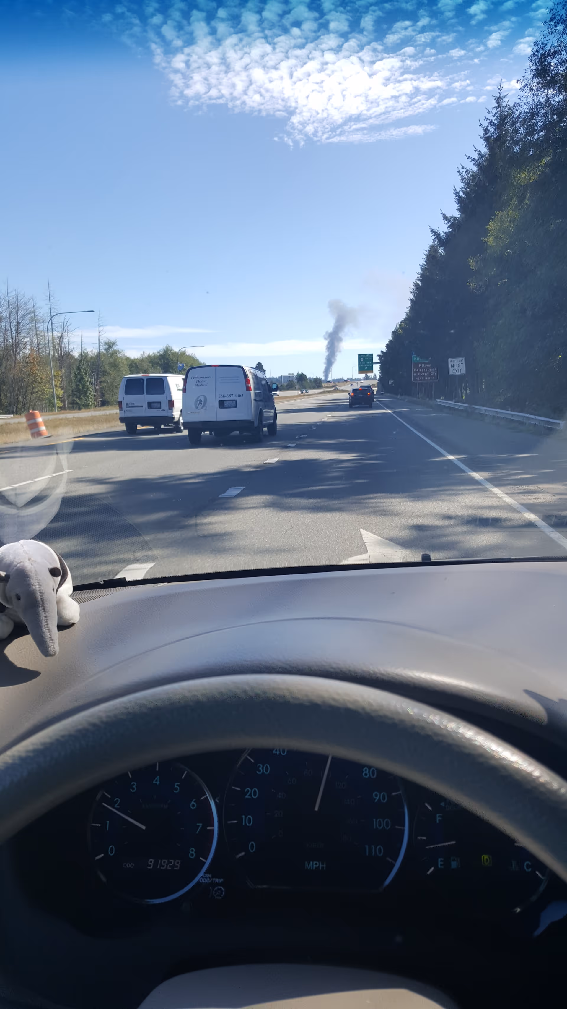 Dashboard view from driver shows highway ahead, smoke plume rising, and a plush toy on dash.