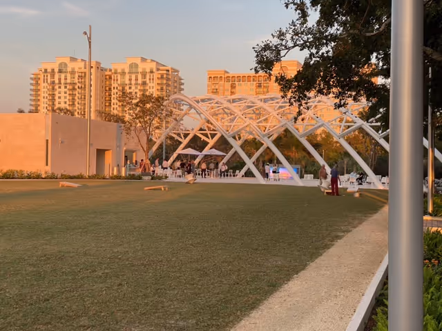 Sunlit outdoor park area with white arched pavilion and surrounding lawn.