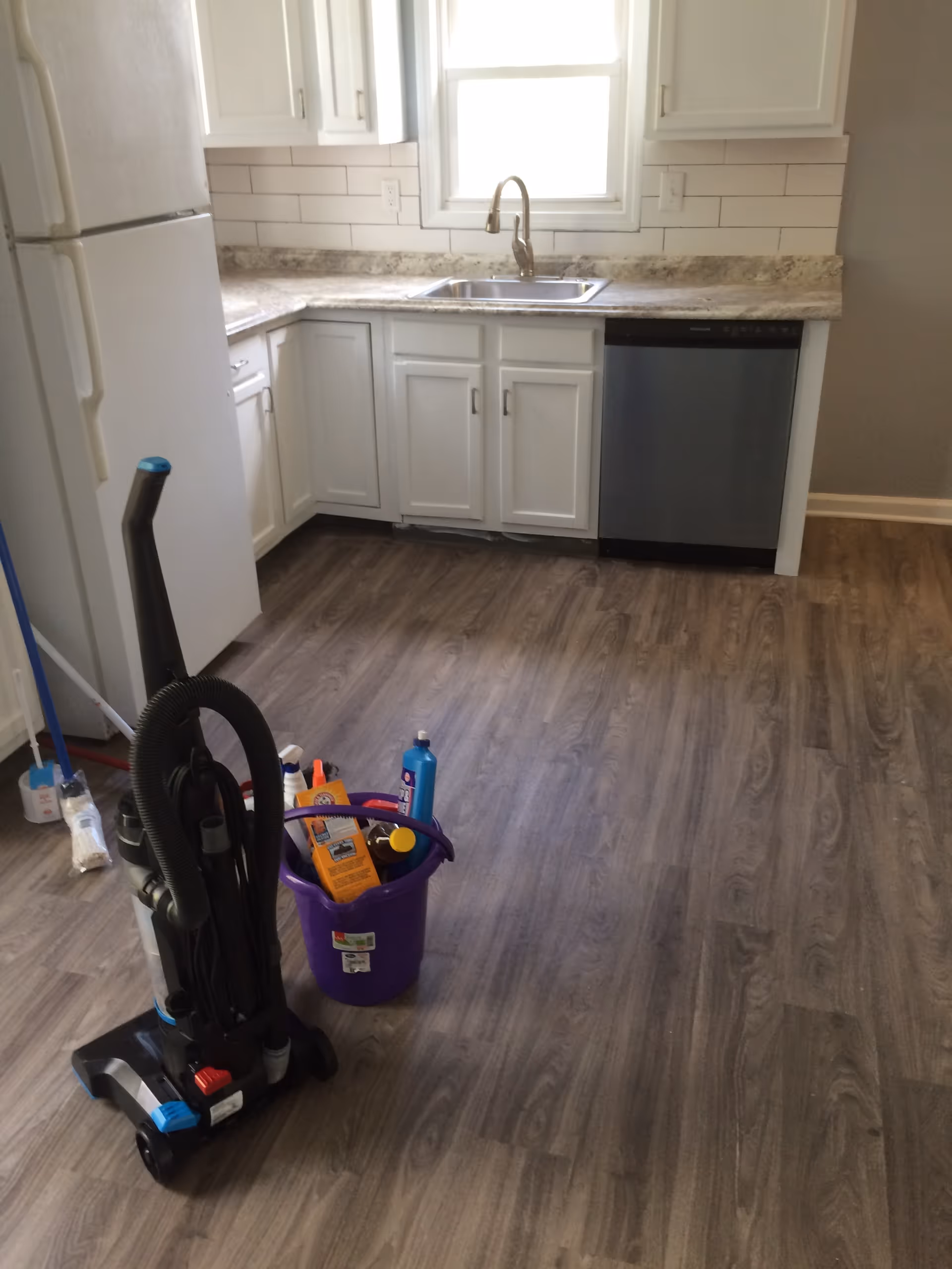 White kitchen with cabinets, sink, dishwasher, and cleaning supplies on wood floor.