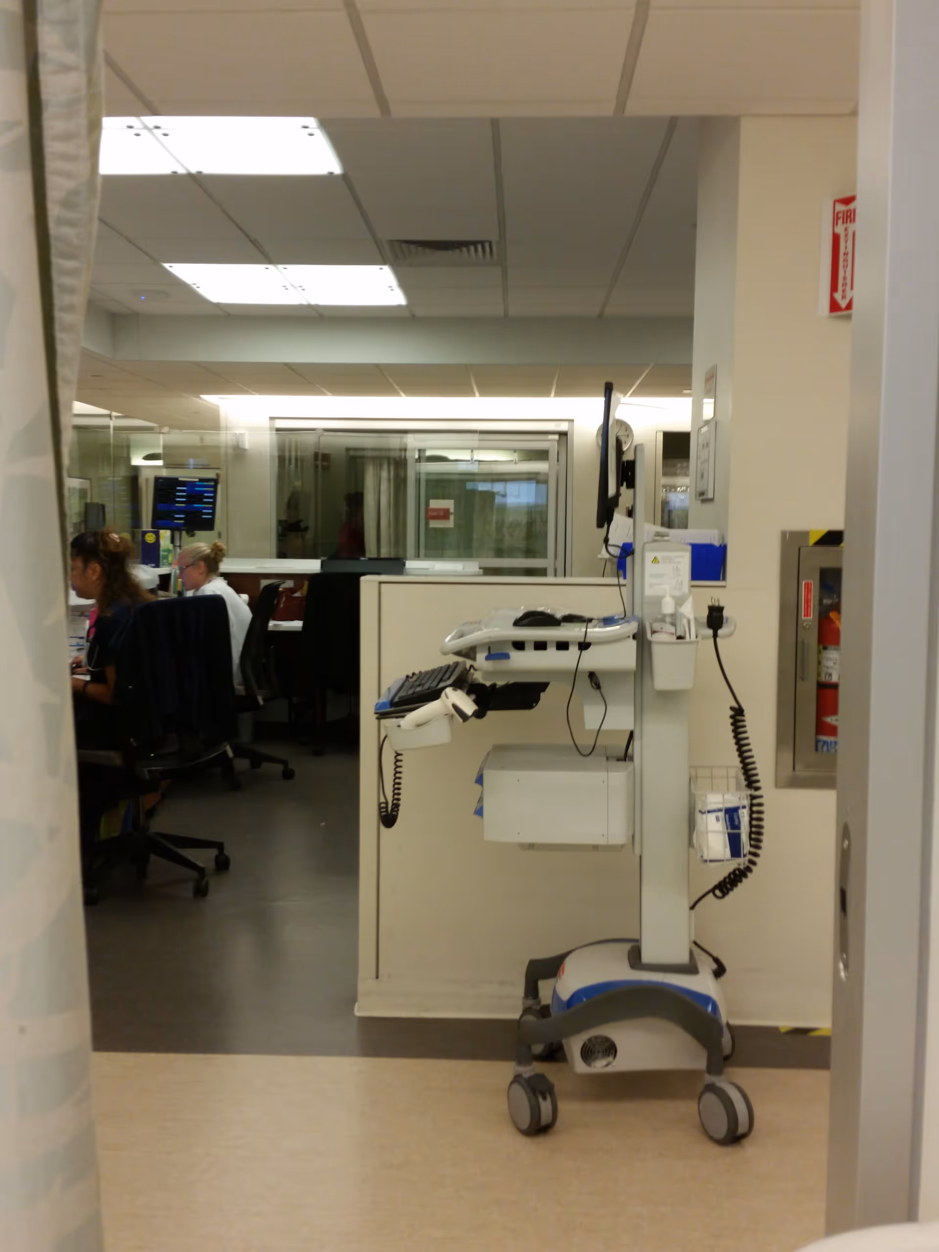A hospital corridor with a mobile medical cart and staff at desks in the background.