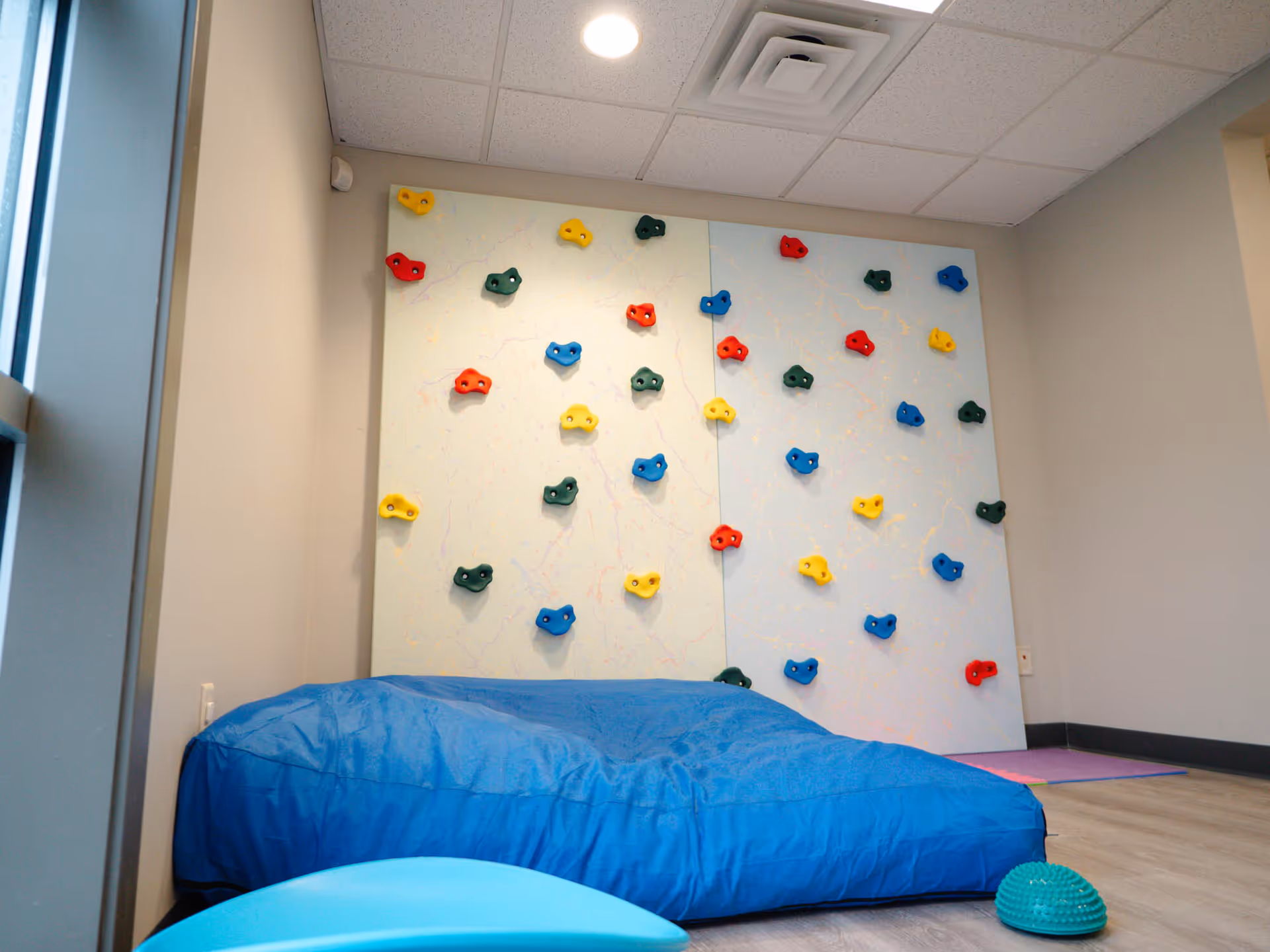 Indoor therapy room with a climbing wall, colorful holds, and blue mats.