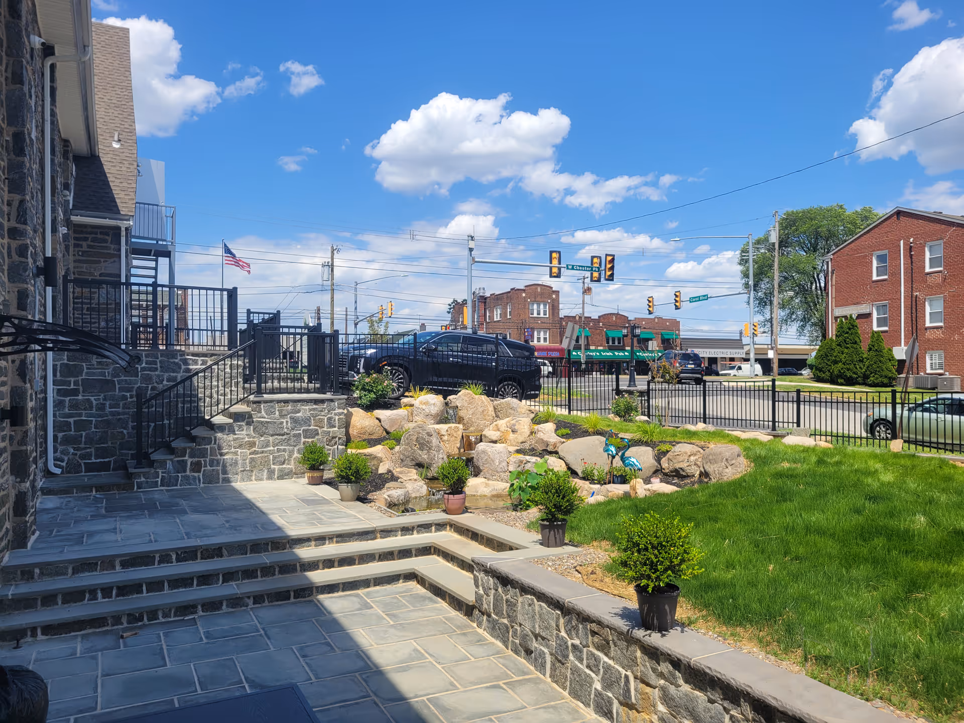 Stone steps, landscaped patio, and potted plants outside a stone-faced facility along a busy street.
