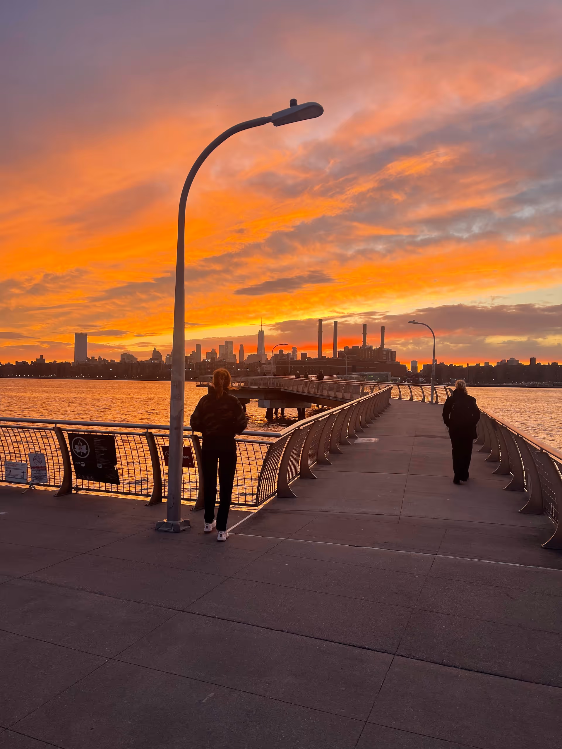 Sunset over a waterfront promenade with two people walking along the curved railing.