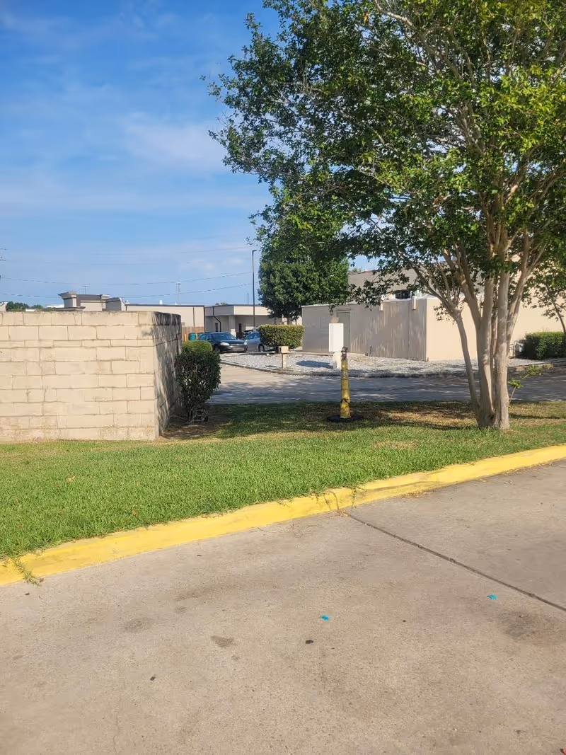 Exterior view of a facility's parking lot with trees, grass, and a low concrete wall.