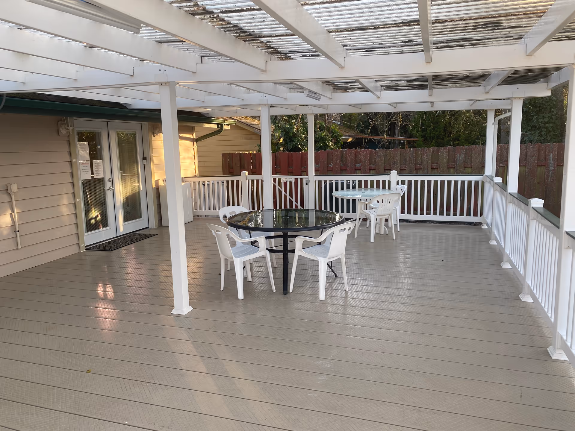 Outdoor deck with glass-topped table, white plastic chairs, and a white pergola.