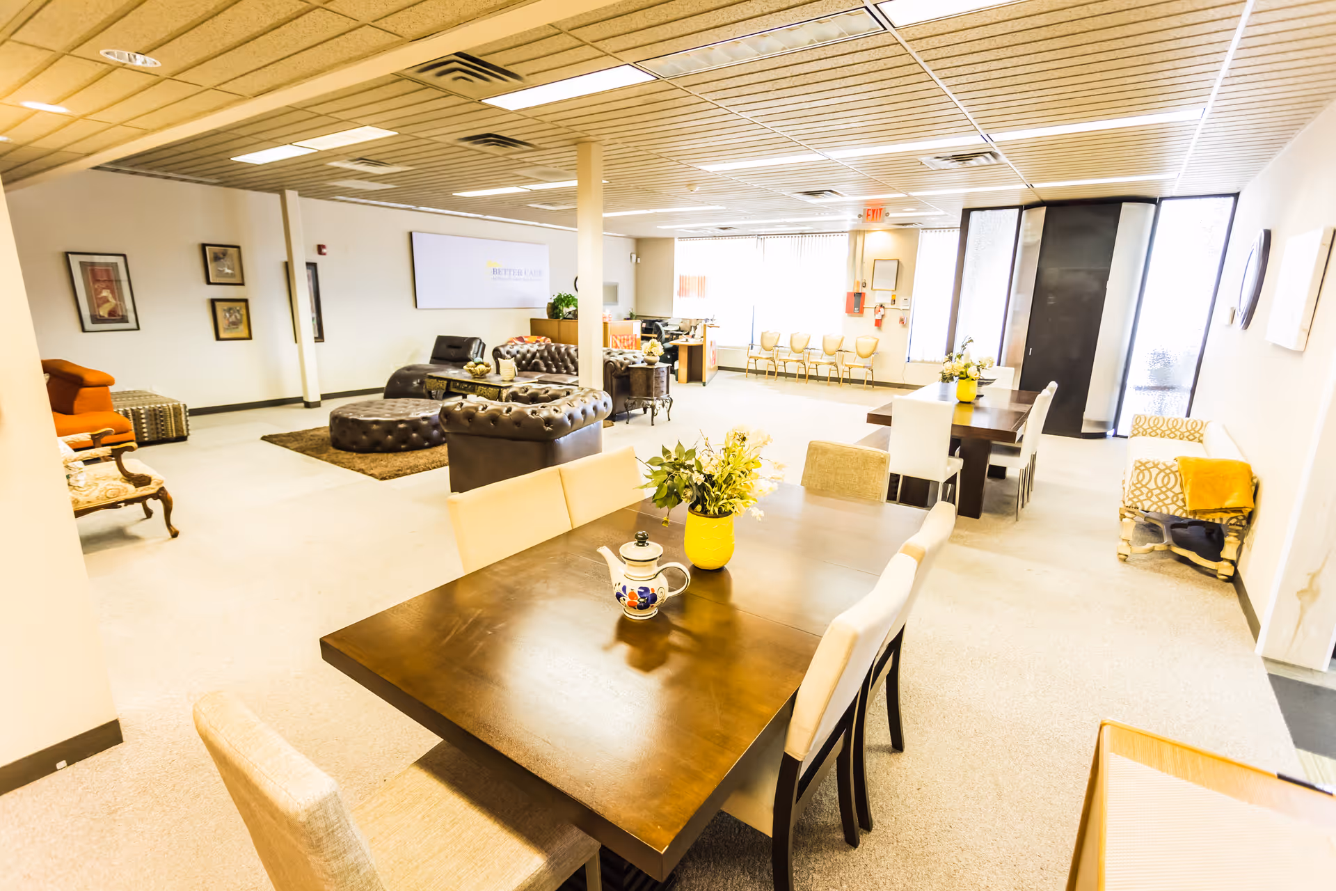 Bright communal lounge with dining tables, sofas, and plants in a senior living facility.