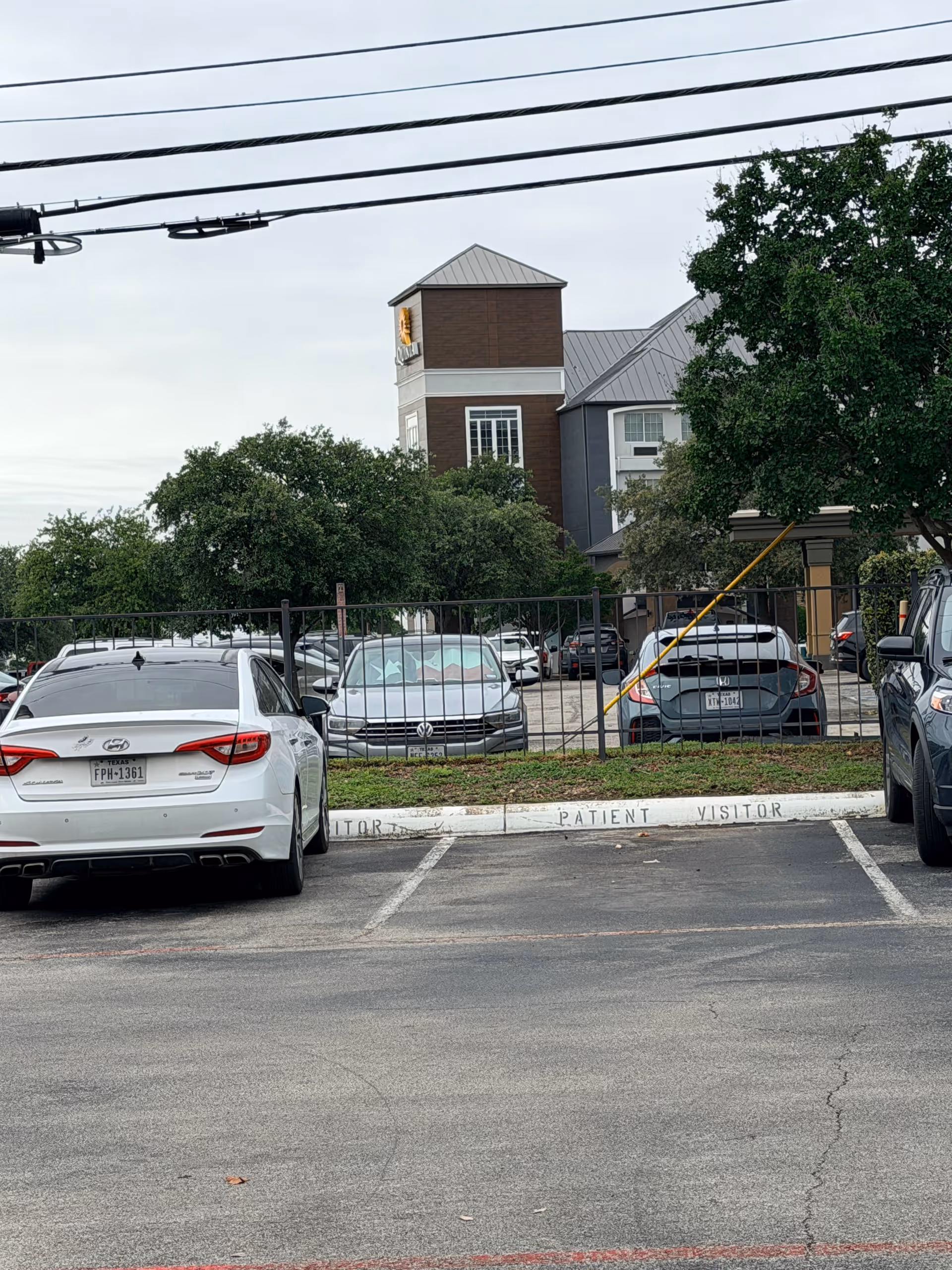 Exterior view of a parking lot in front of a brick building with trees and fence.