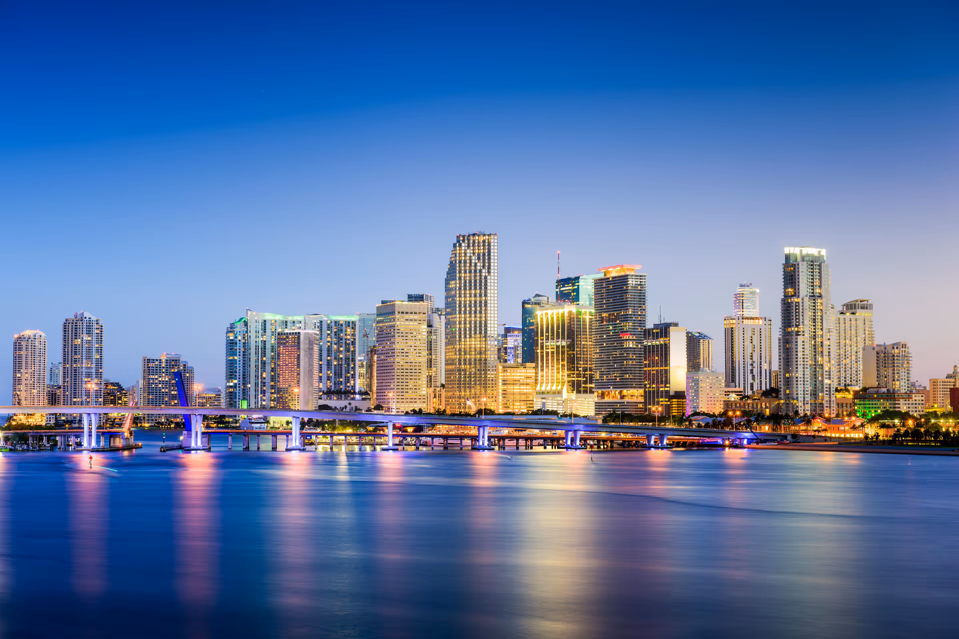 Dusk city skyline across a calm river with illuminated high-rise buildings.
