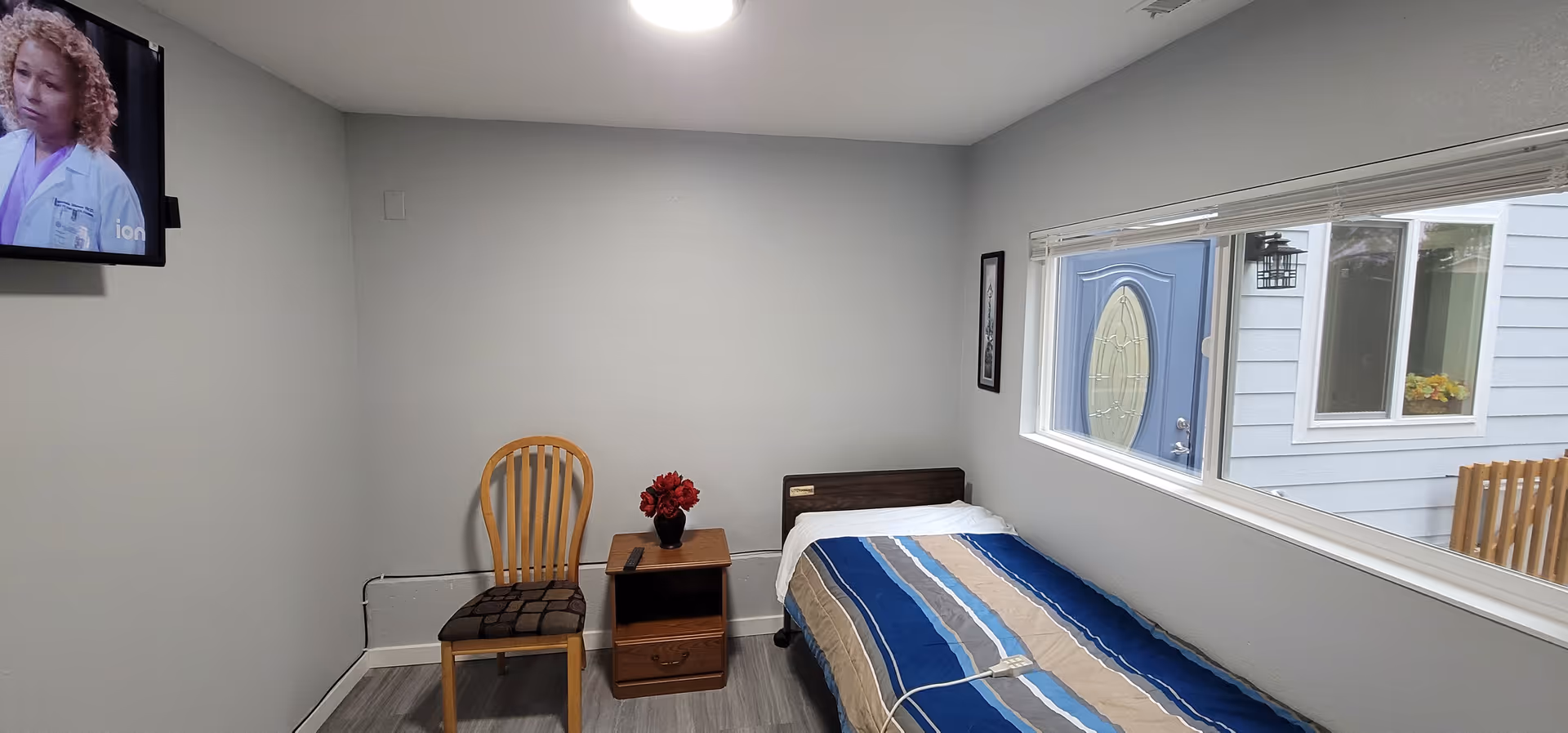 Sparse bedroom in a senior facility with a single bed, chair, nightstand, and TV.