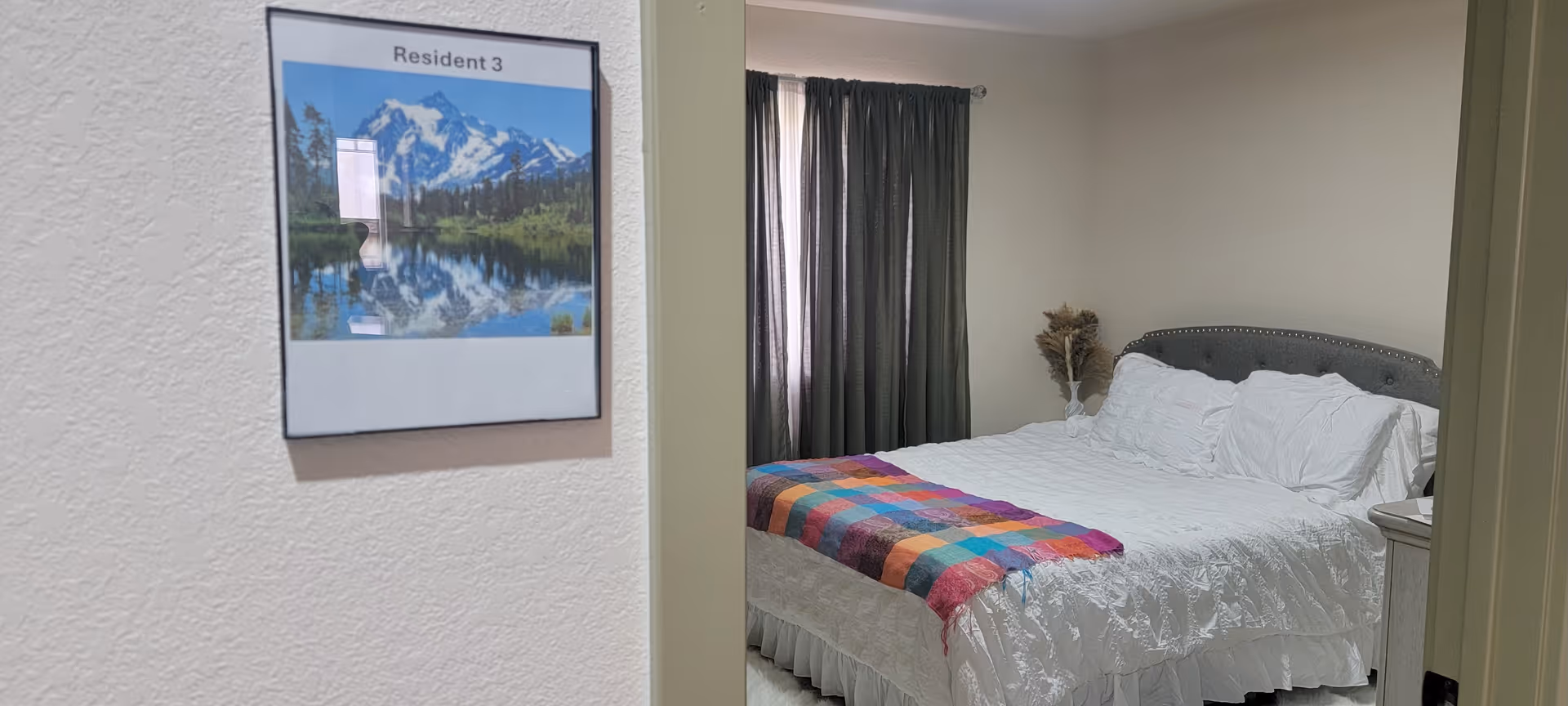Neatly made bedroom in a senior living facility with framed art and dark-curtained window.