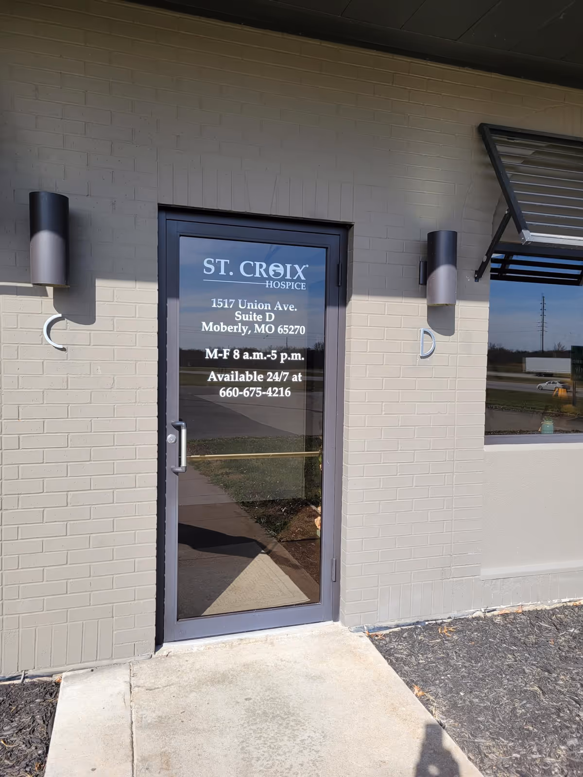 Glass door entrance of St. Croix Hospice with address and hours posted.