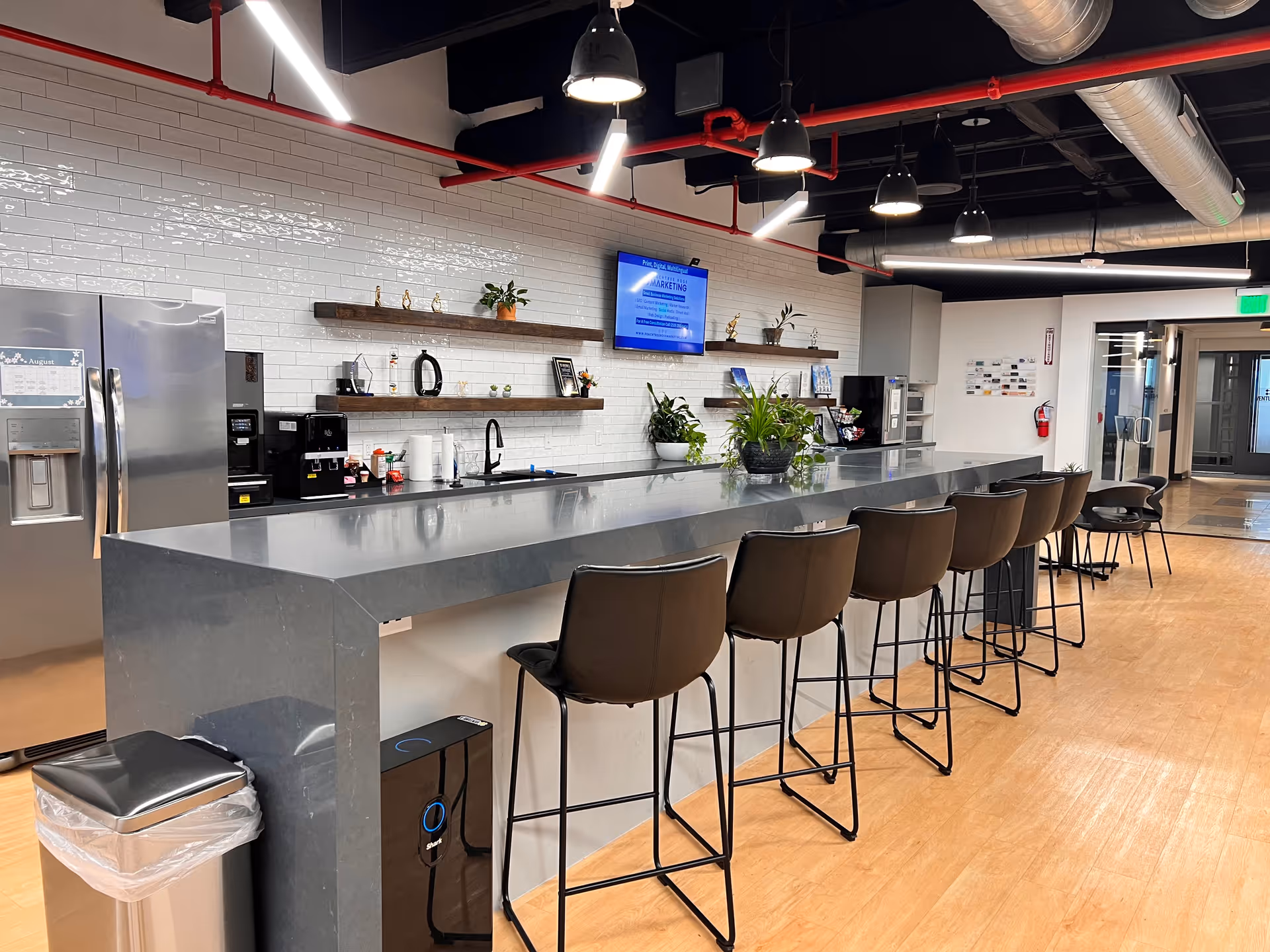 Long modern service counter with brown stools, white tiled wall, shelves, plants, and a coffee area.
