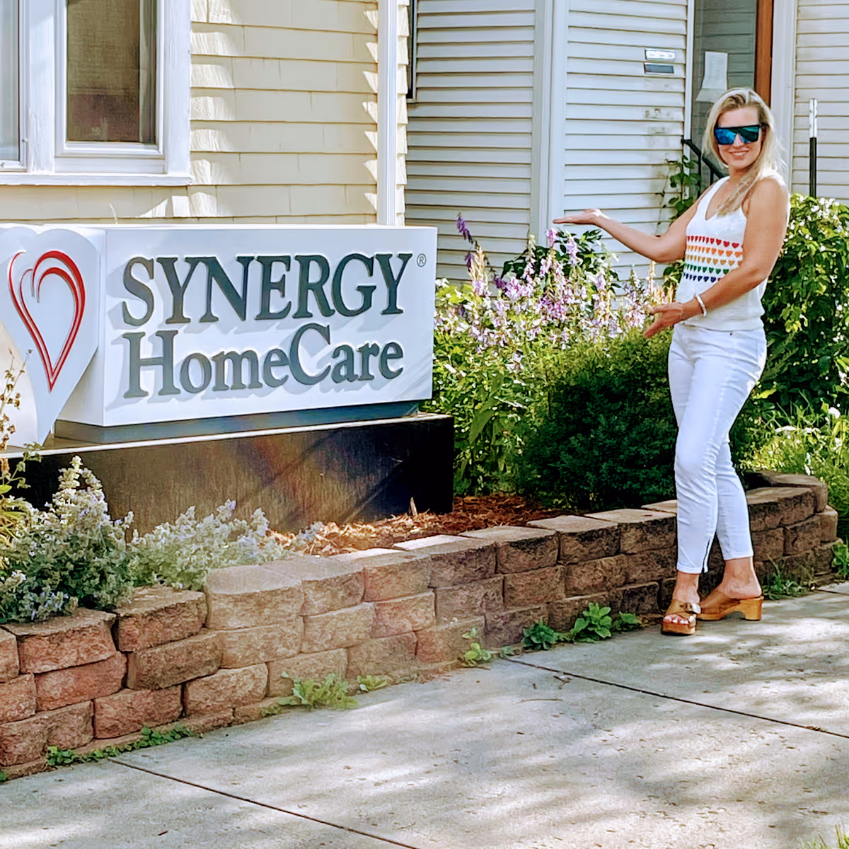 A woman stands beside a SYNERGY HomeCare sign outside a residential building exterior.