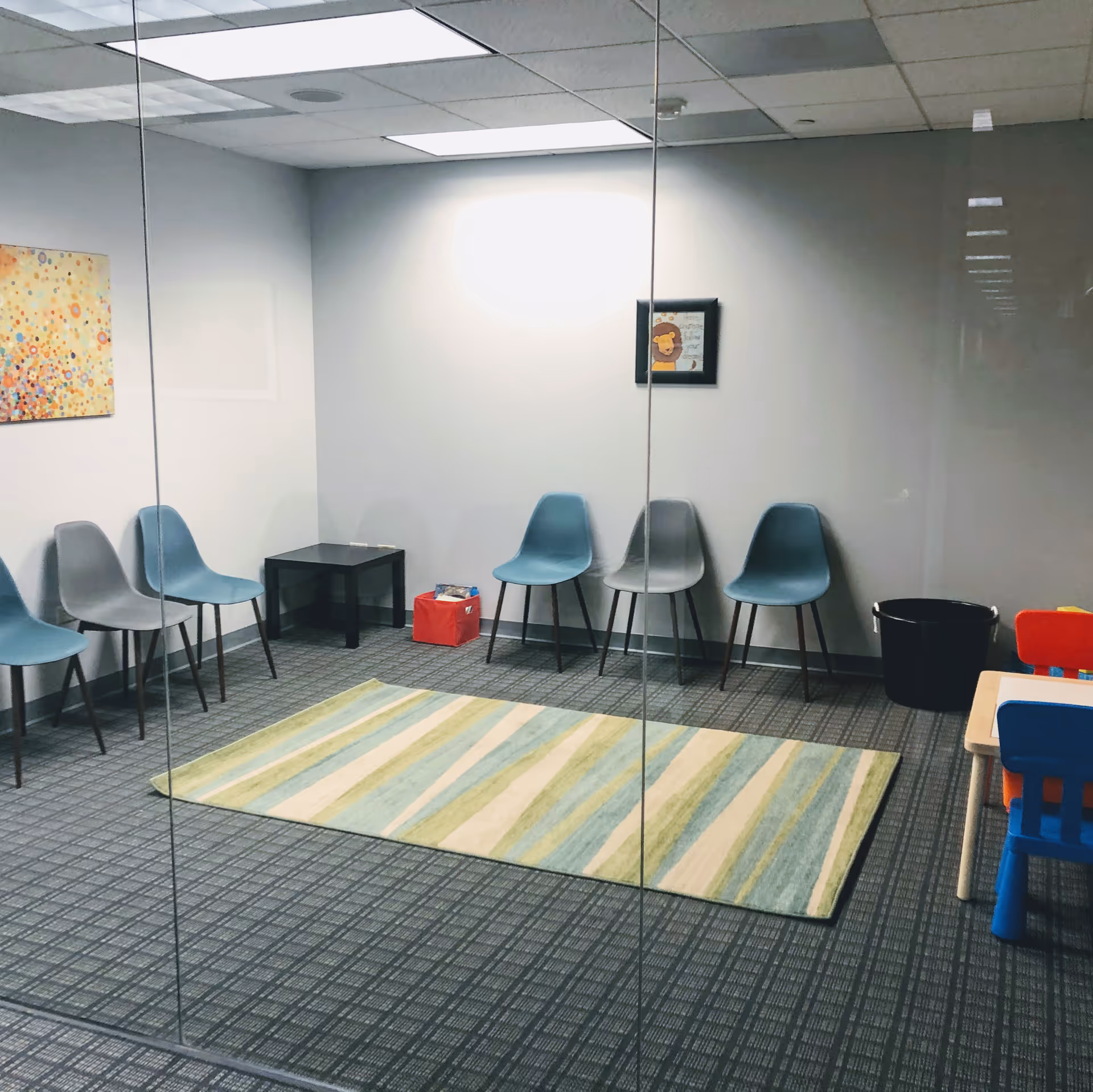Glass-walled waiting room with blue chairs, rug, and children's play area.