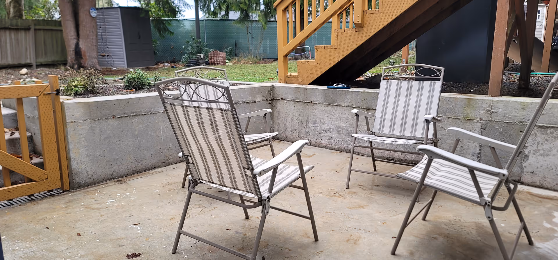 Outdoor concrete patio with striped chairs and a wooden staircase in the background.