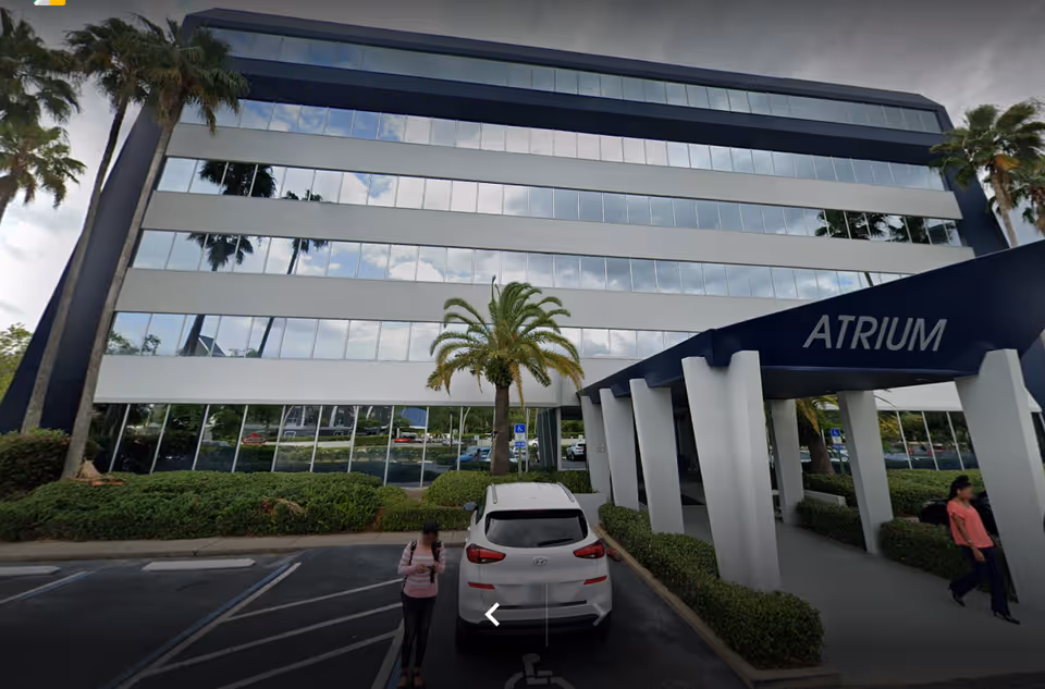 Exterior of a modern glass-front building with palm trees and a central atrium entrance.