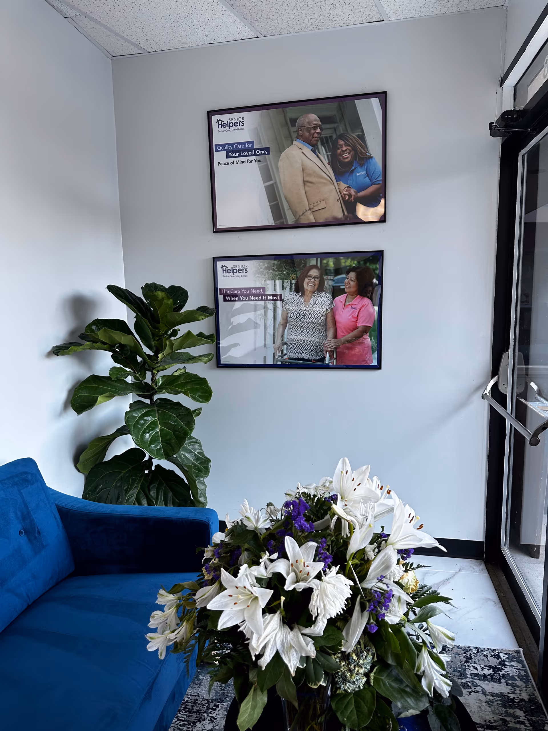 Lobby seating area with blue sofa, potted plant, flower arrangement, and framed Senior Helpers posters.