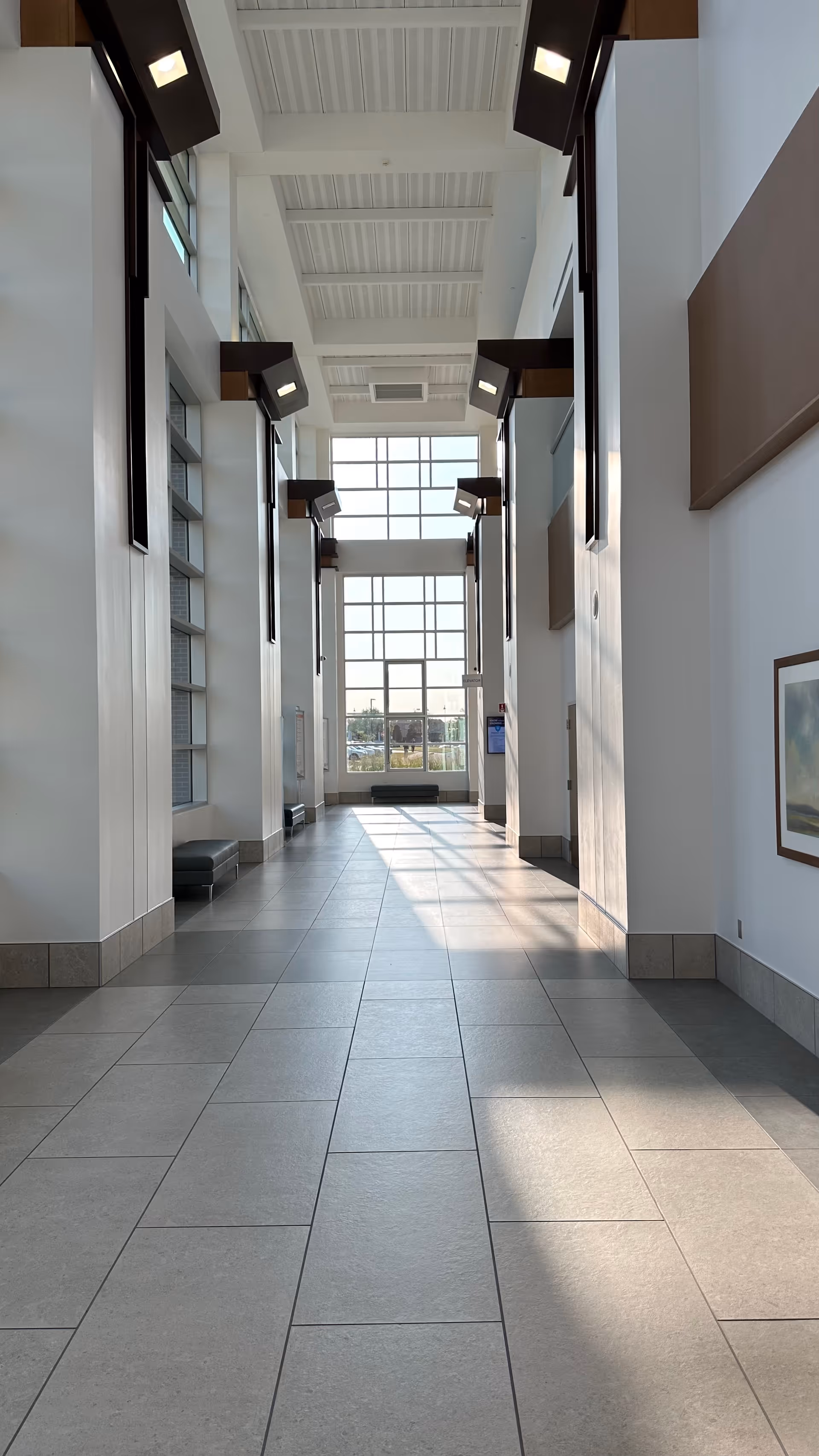 Bright, spacious interior hallway with large windows, tiled floor, and wall-mounted light fixtures.
