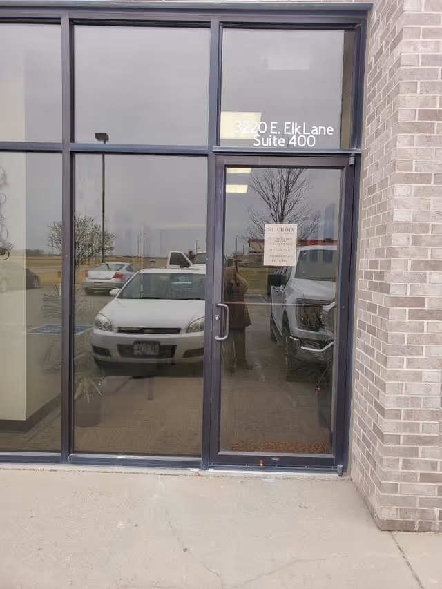 Glass entrance doors of St. Croix Hospice building with address sign and parked cars reflected