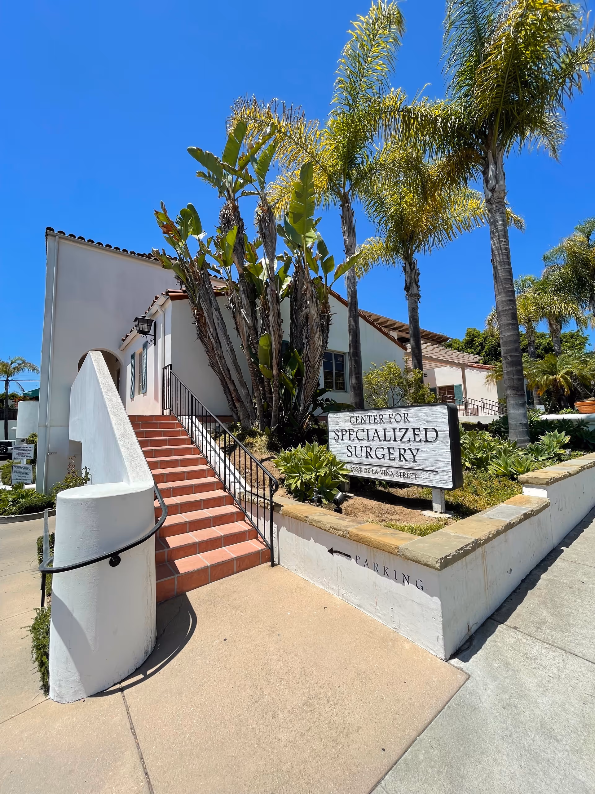 White stucco building with red-tiled stairs, tropical plants, and a sign reading Center for Specialized Surgery.