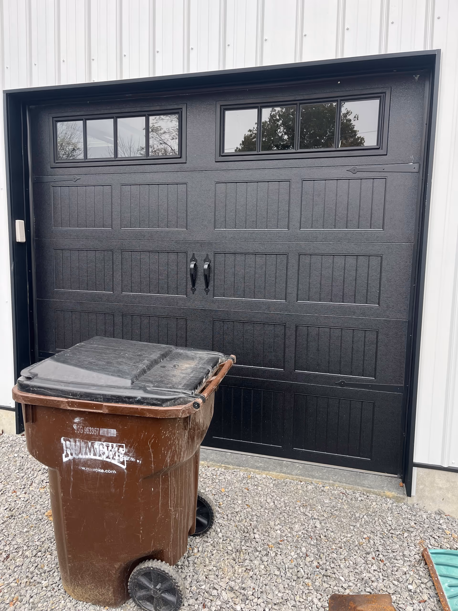 Dark garage-style door on a light metal building facade with a brown wheeled trash bin.