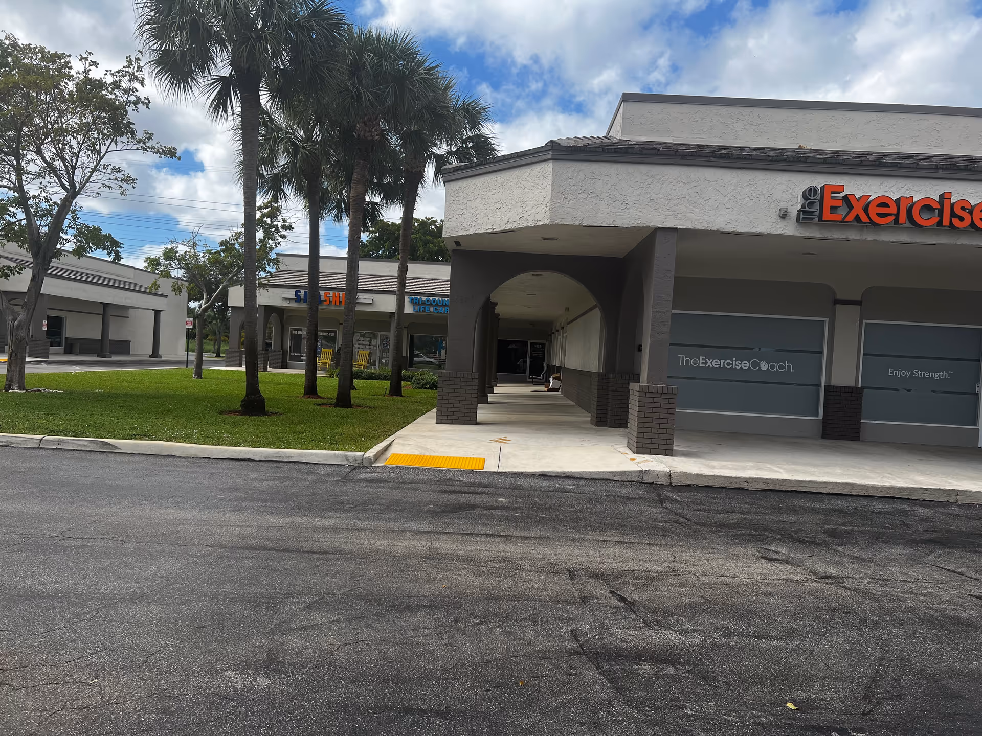 Exterior view of a strip mall-style facility with palm trees, walkway, and storefronts.