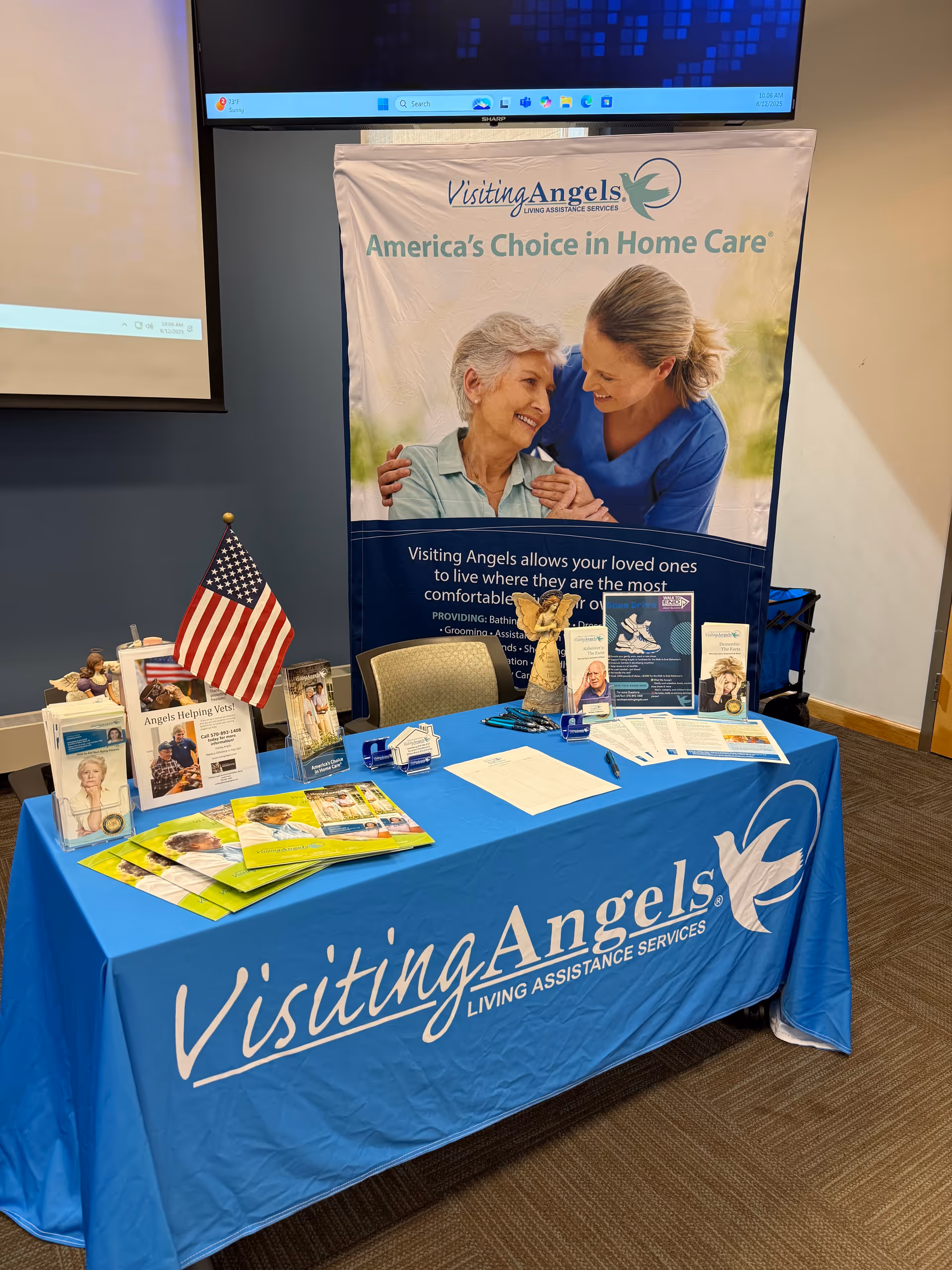 Indoor Visiting Angels promotional booth with banner, table, brochures, and an American flag.