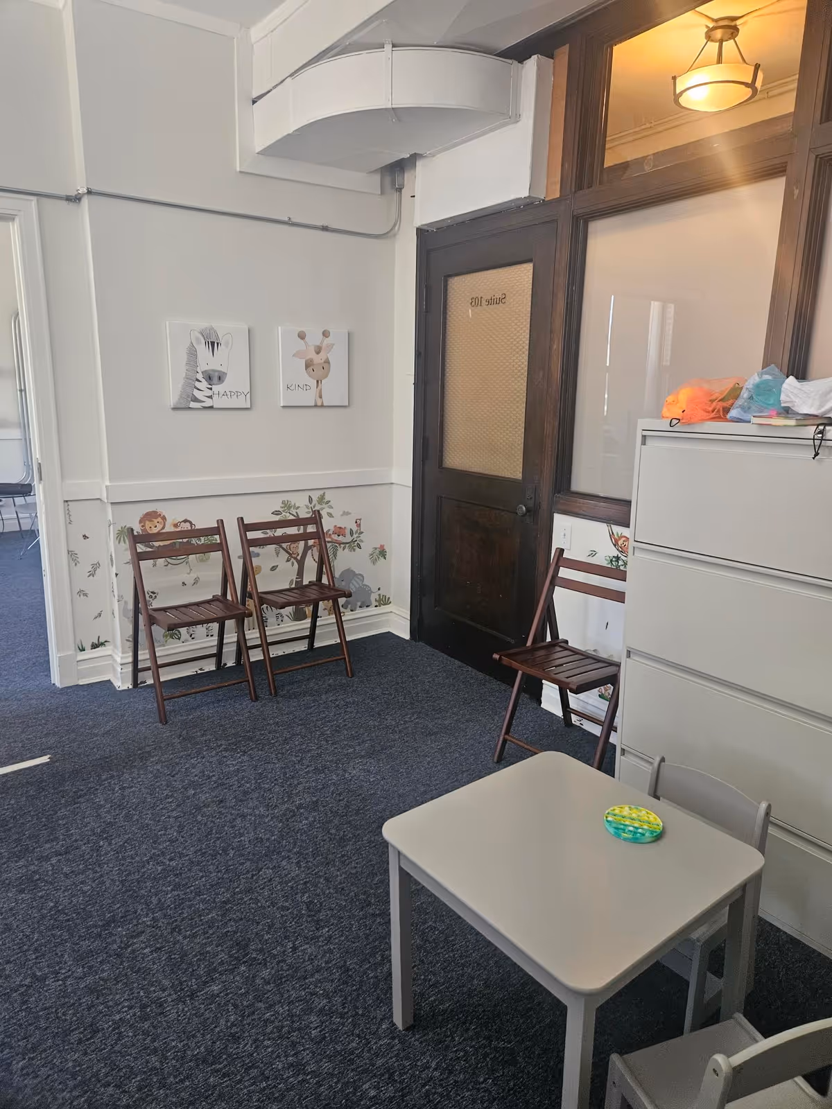 Interior common room with small table, wooden chairs, a storage cabinet, and decorative wall art.
