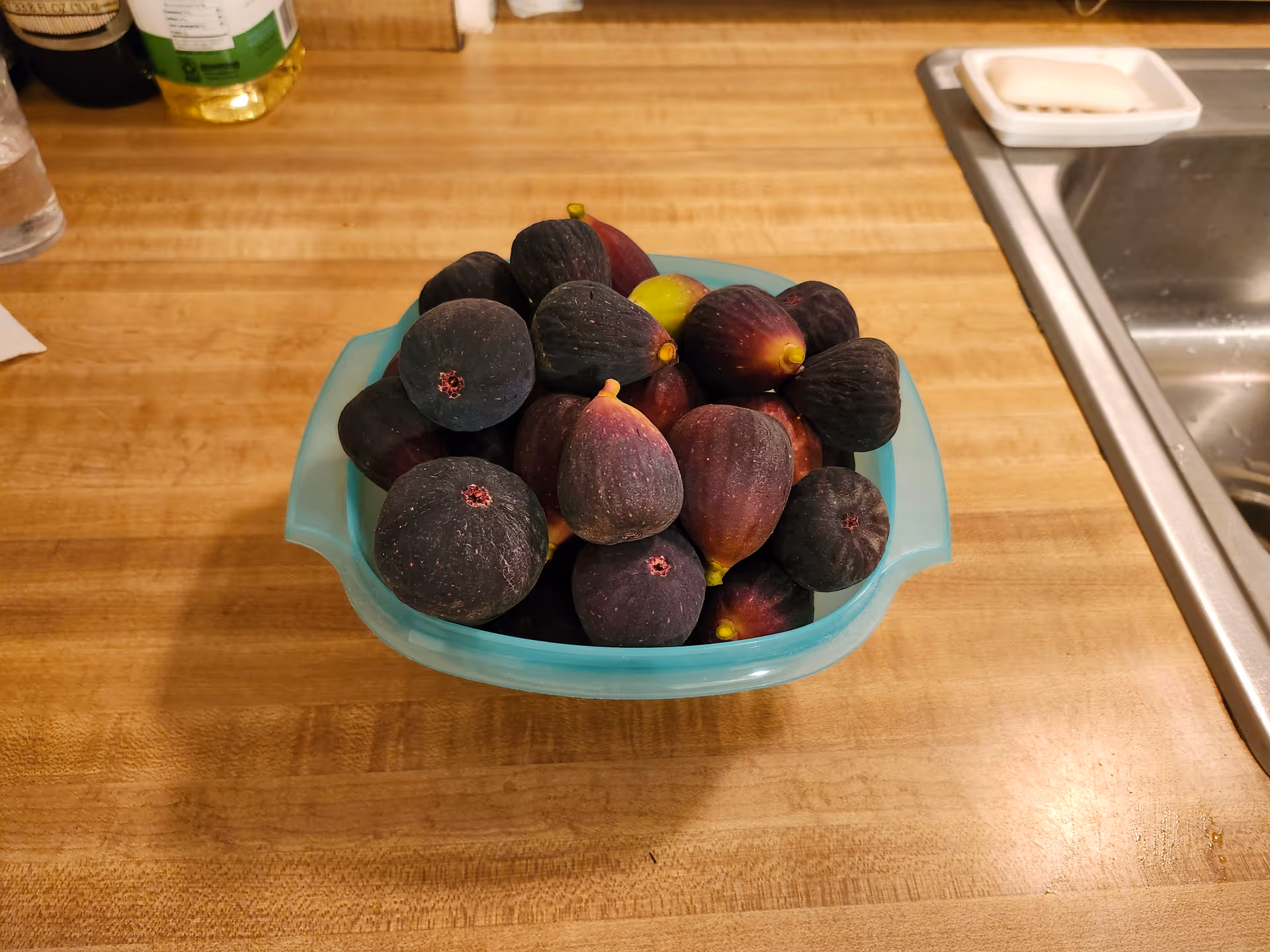 Turquoise bowl of ripe figs sits on a wooden kitchen counter beside a sink.