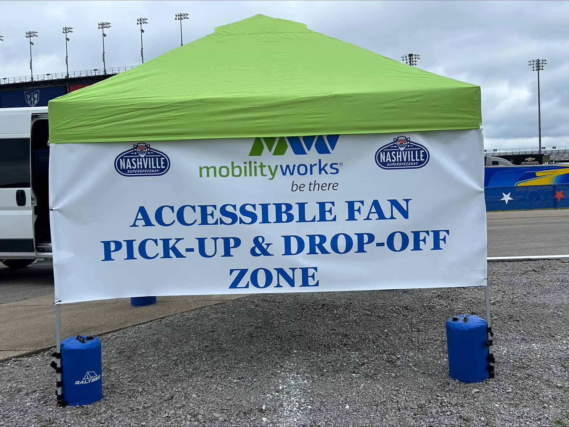 Green tent with banner reading Accessible Fan Pick-Up & Drop-Off Zone at a race track.