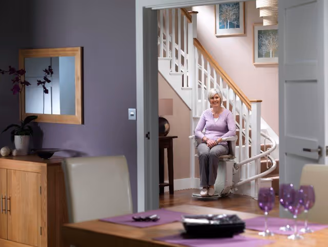 Elderly woman seated in a stair lift at a staircase inside a dining area.