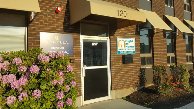 Brick building front with entrance door, Right at Home sign, awnings, and flowering shrubs.